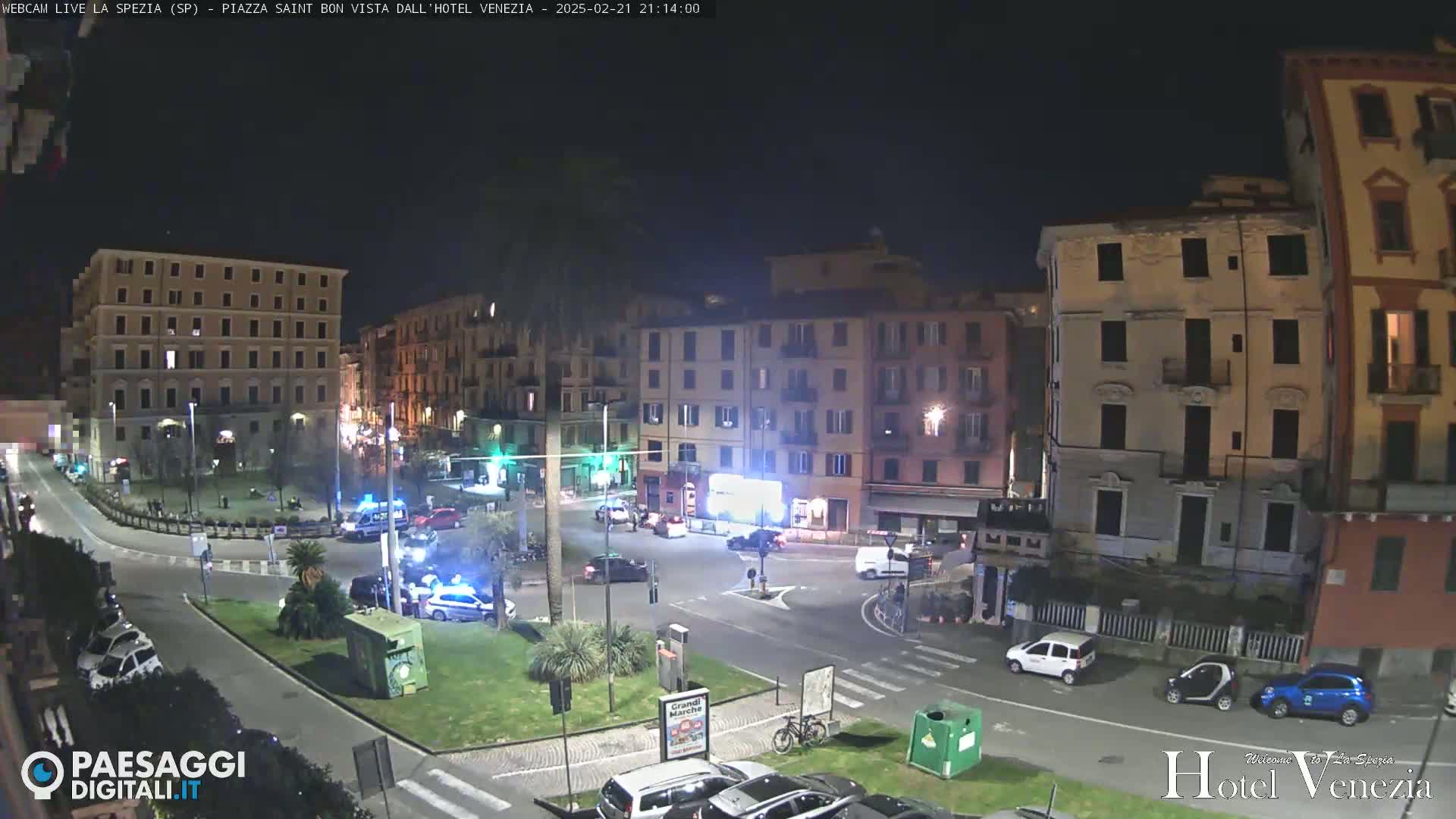 A nighttime view of a city square with several buildings, cars, and a few pedestrians, under clear skies.