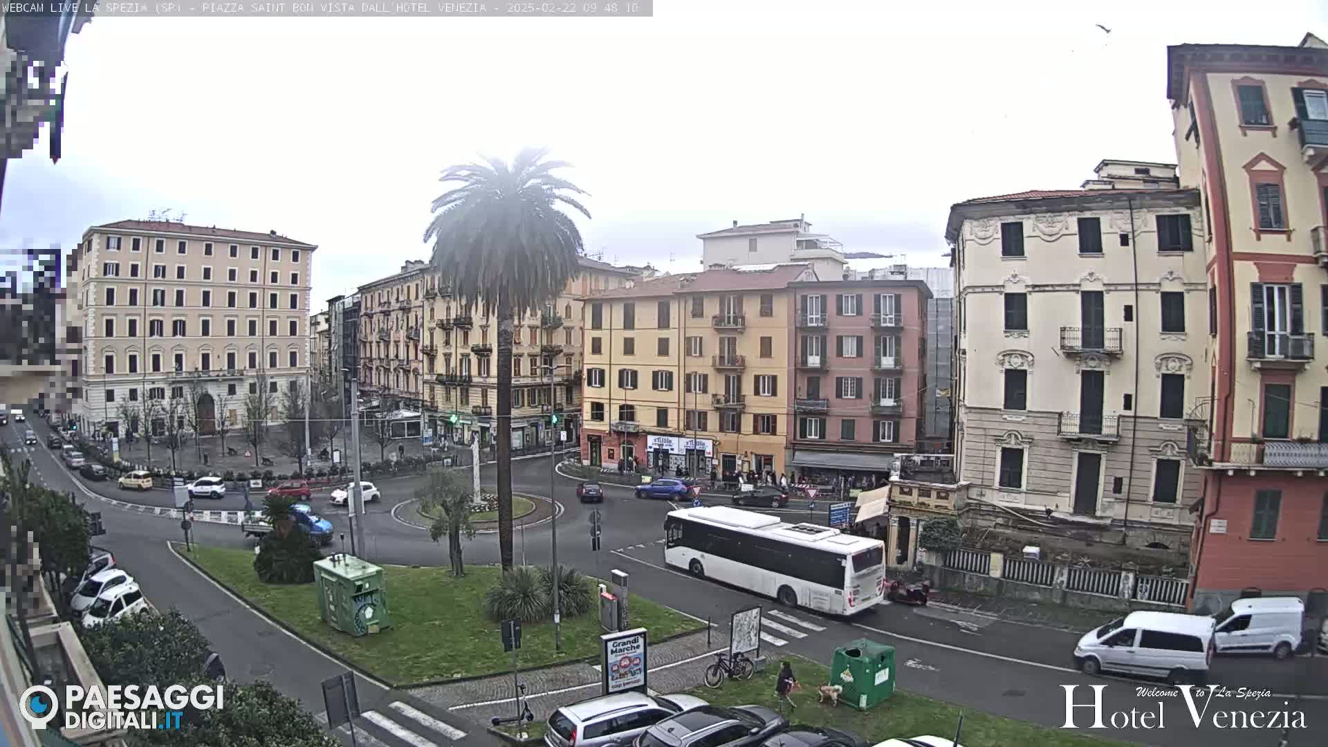 A mostly cloudy day reveals a city square with several multi-story buildings, a roundabout, parked cars, a bus, and a lone palm tree at the center.