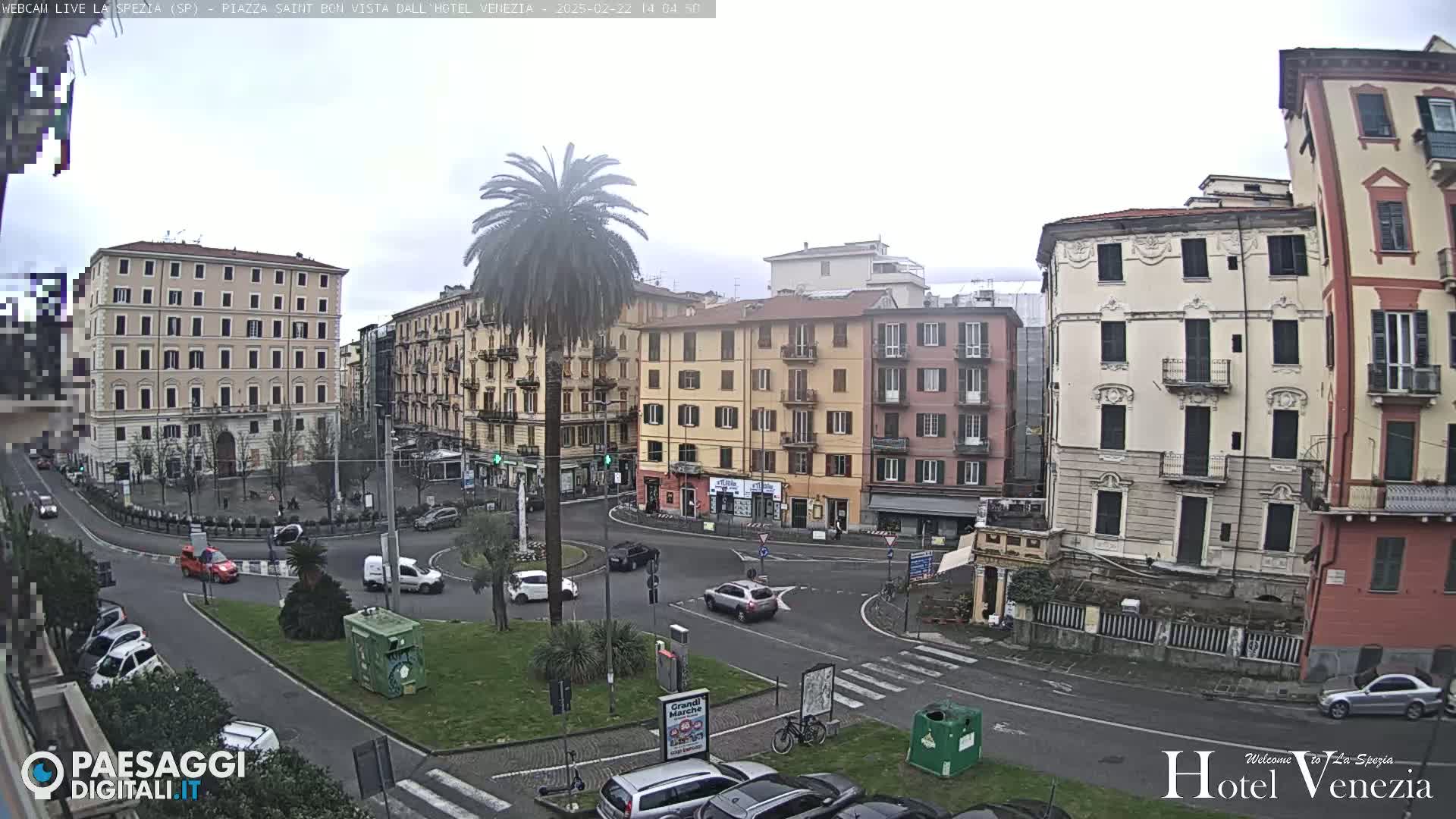 A mostly overcast day reveals a European city square with several multi-story buildings surrounding a roundabout, with cars and a lone palm tree visible.