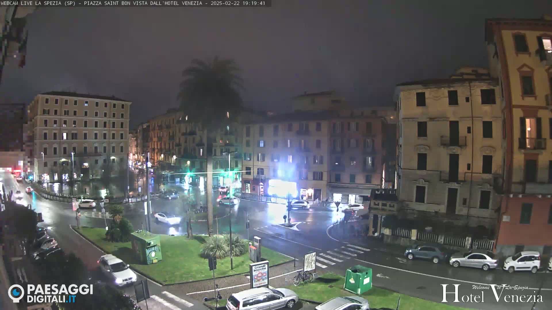 A nighttime, rain-slicked city square is surrounded by multi-story buildings, with a few cars visible on the wet roads.
