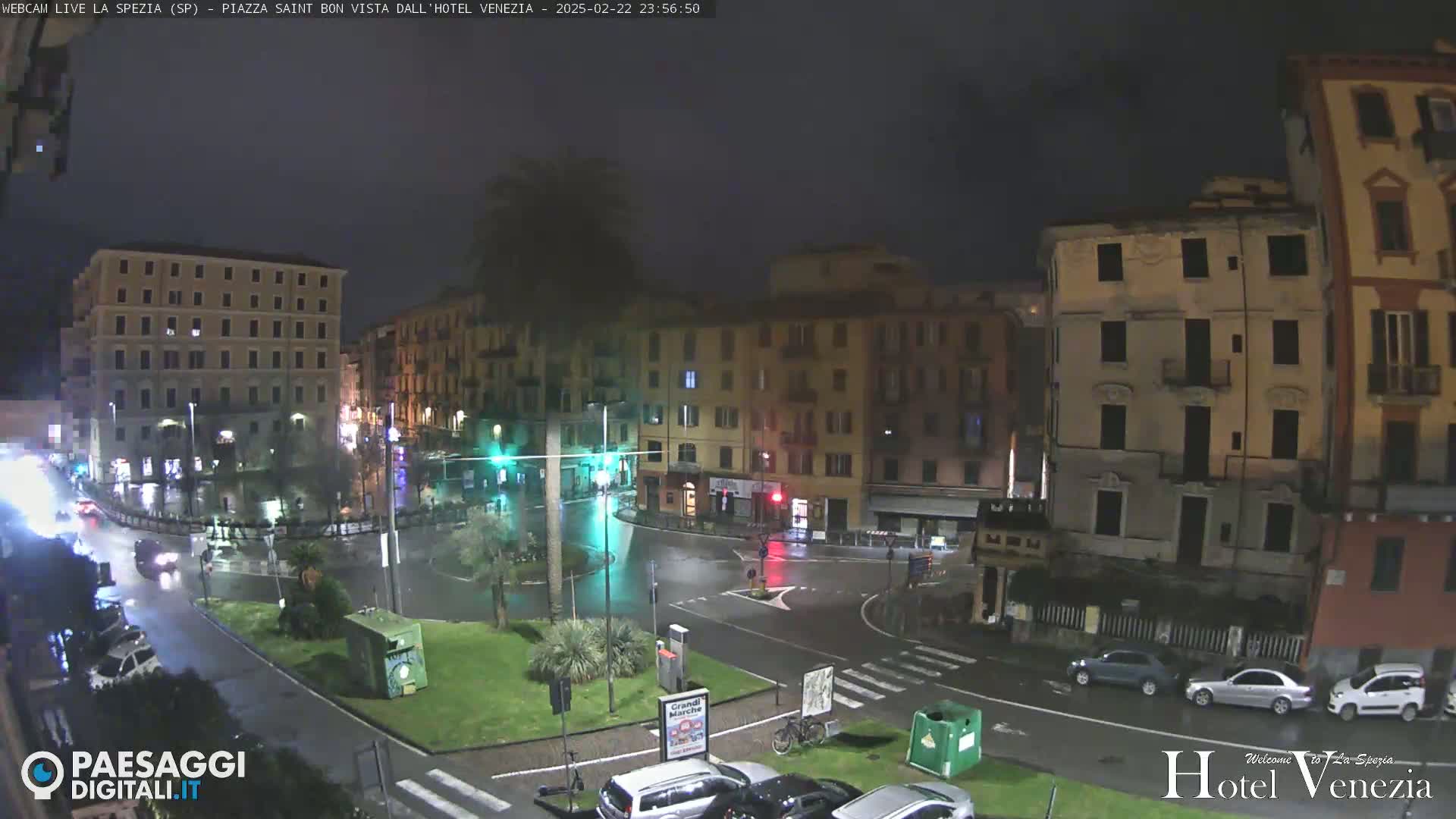 A rain-slicked city square at night, showing several multi-story buildings and sparse traffic.