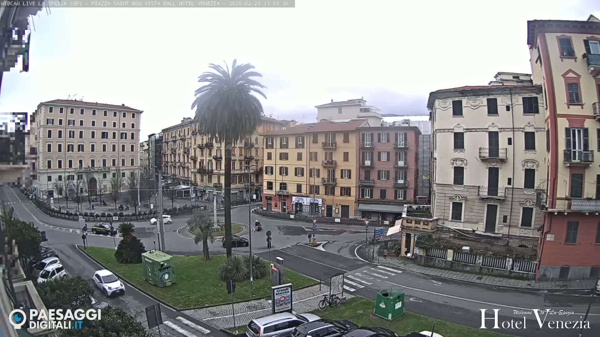 A city square with several multi-story buildings, a central roundabout, and a palm tree is visible on an overcast day.