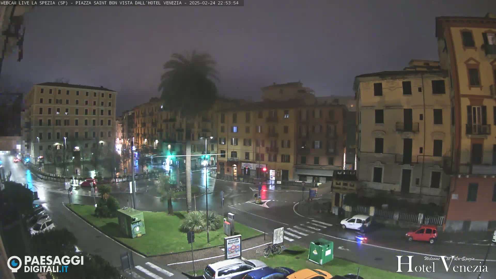 A rain-slicked city square at night, surrounded by multi-story buildings, with a few cars and a palm tree visible.