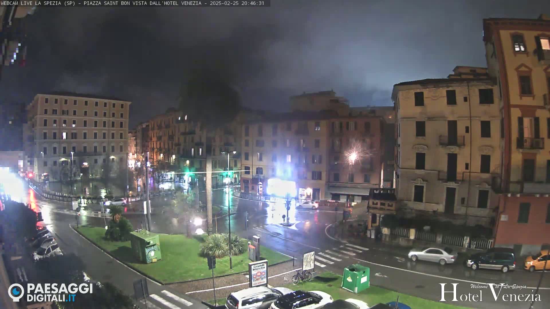 A nighttime, rainy scene of a city square with several multi-story buildings, cars, and a small central park.
