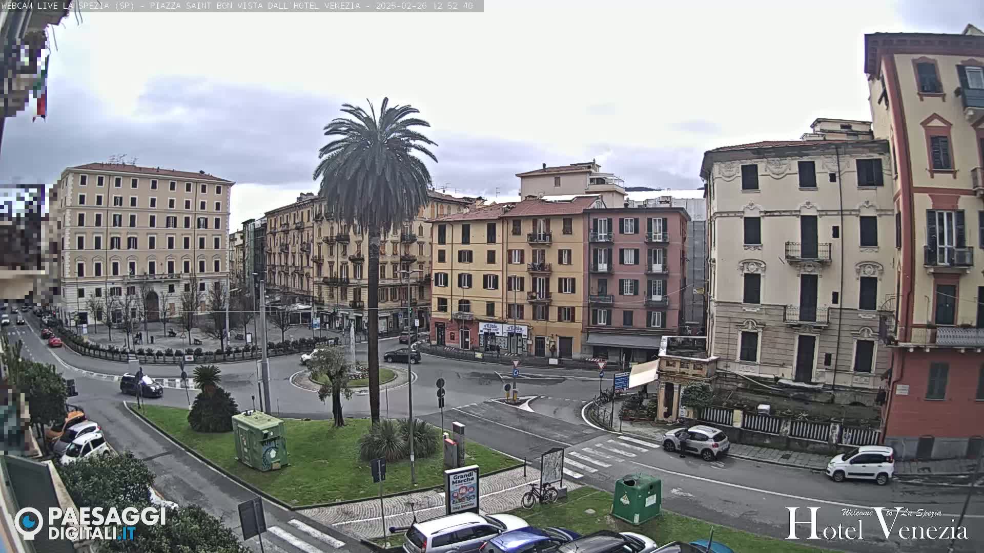 A mostly cloudy day reveals a city square with several multi-story buildings, a central palm tree, and cars parked along the perimeter; a few cars are driving through the square.