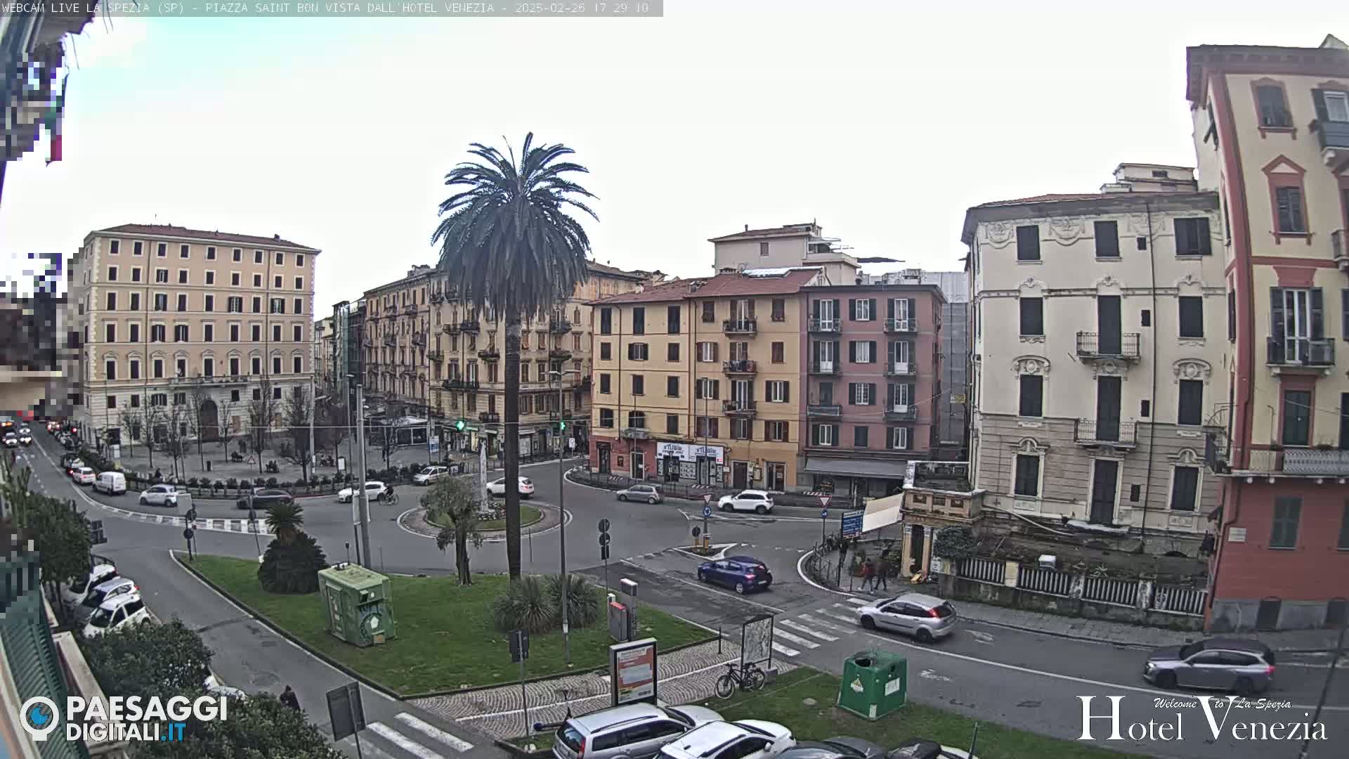 A mostly cloudy day shows a view of a city square with several multi-story buildings, a palm tree in the center, and cars circulating around it.