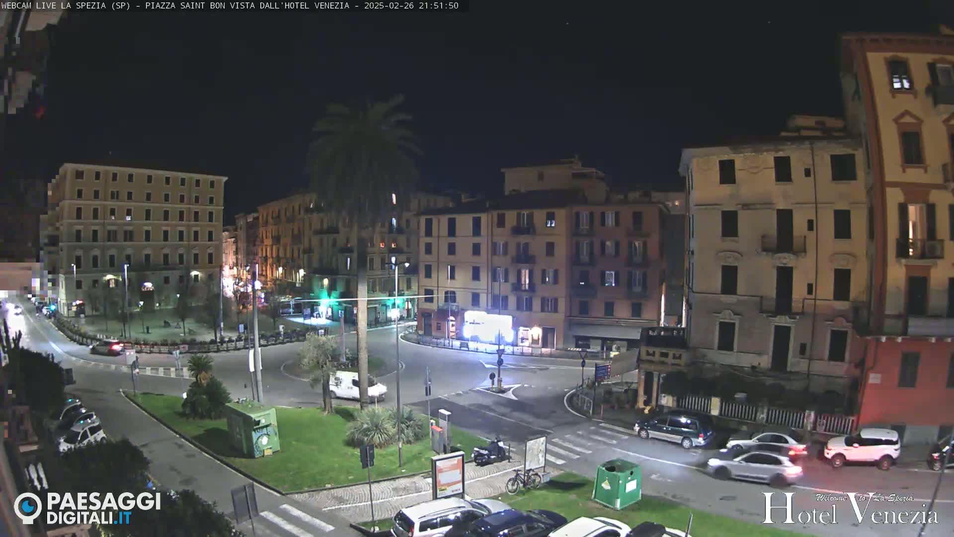 A nighttime view of a European city square with several buildings surrounding a roundabout, where cars are driving and a few pedestrians are visible under a clear night sky.