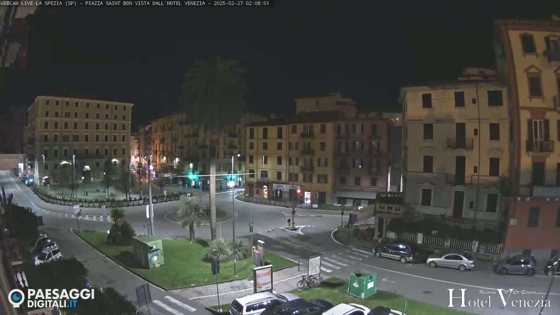 A nighttime view of a town square with several multi-story buildings surrounding a circular roadway, illuminated by streetlights.