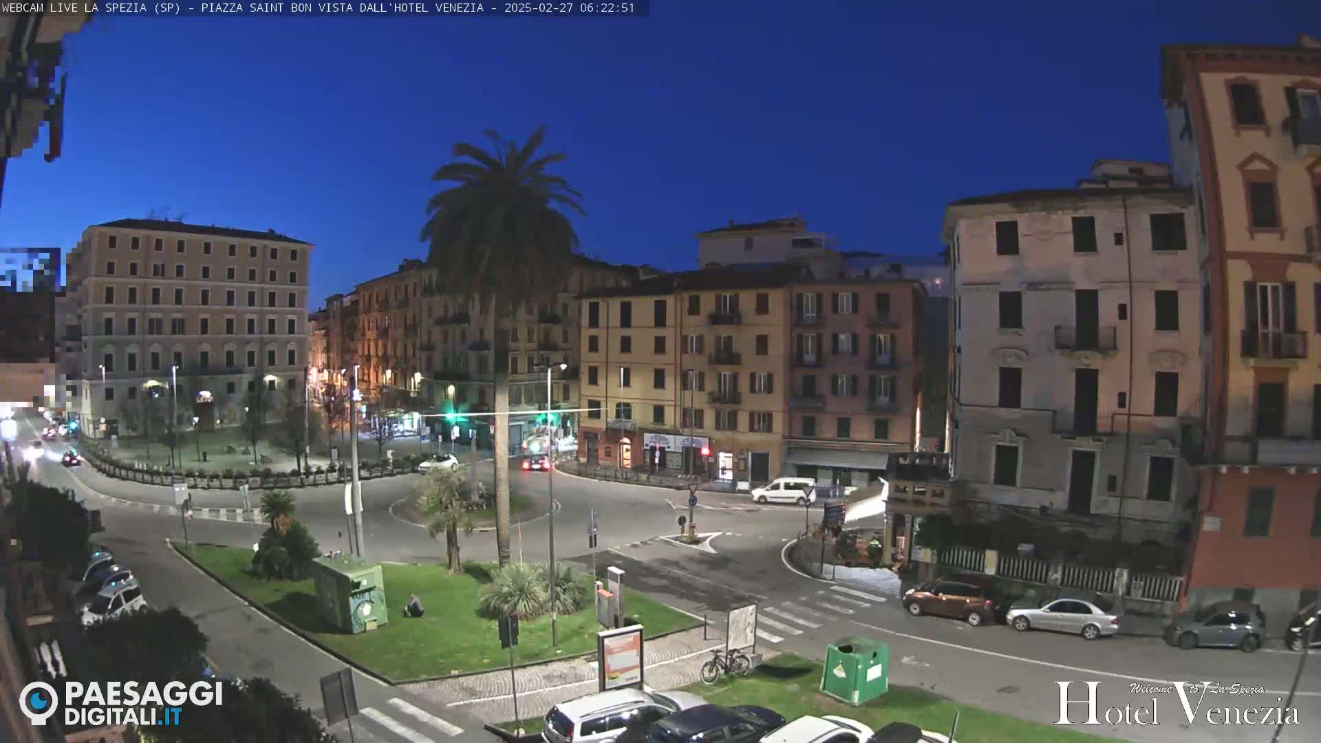A twilight view of a city square with cars parked and driving around a roundabout, surrounded by multi-story buildings under a clear, dark blue sky.