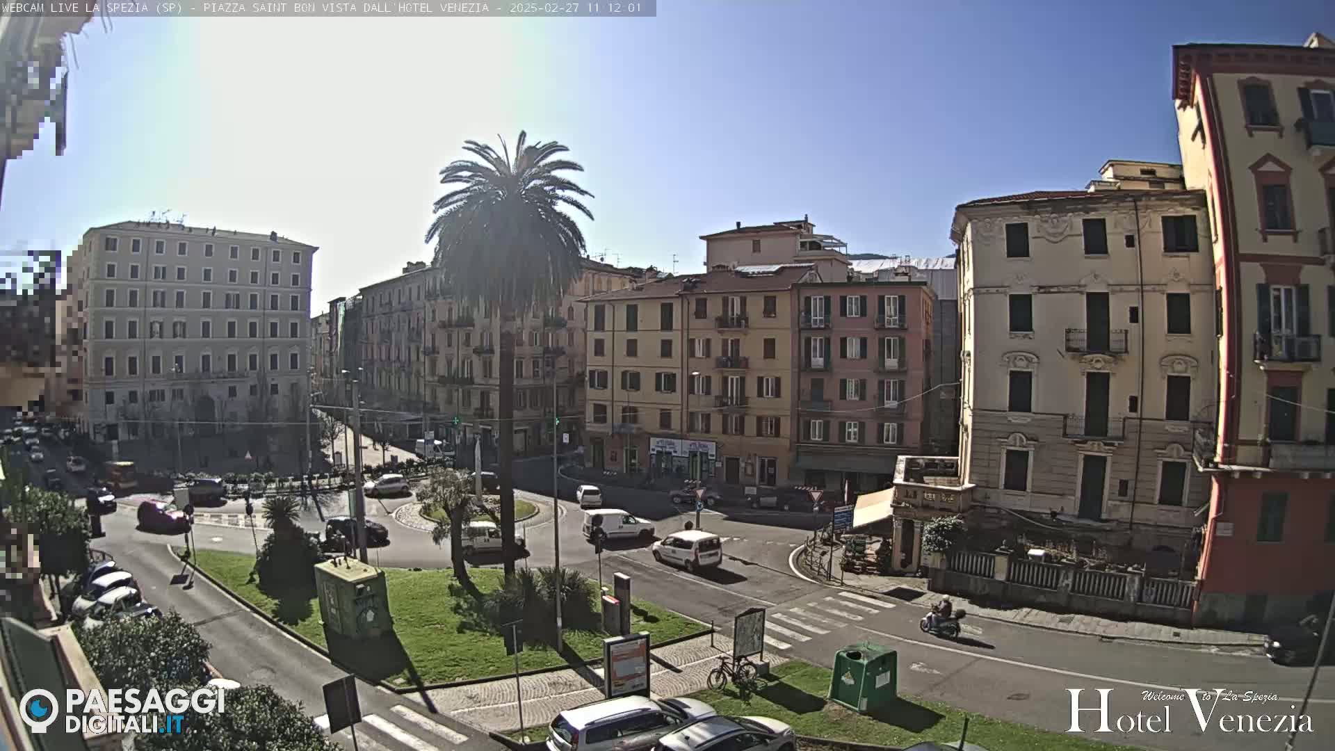 A sunny day in a European city square, showing several multi-story buildings surrounding a traffic circle with cars and a palm tree in the center.