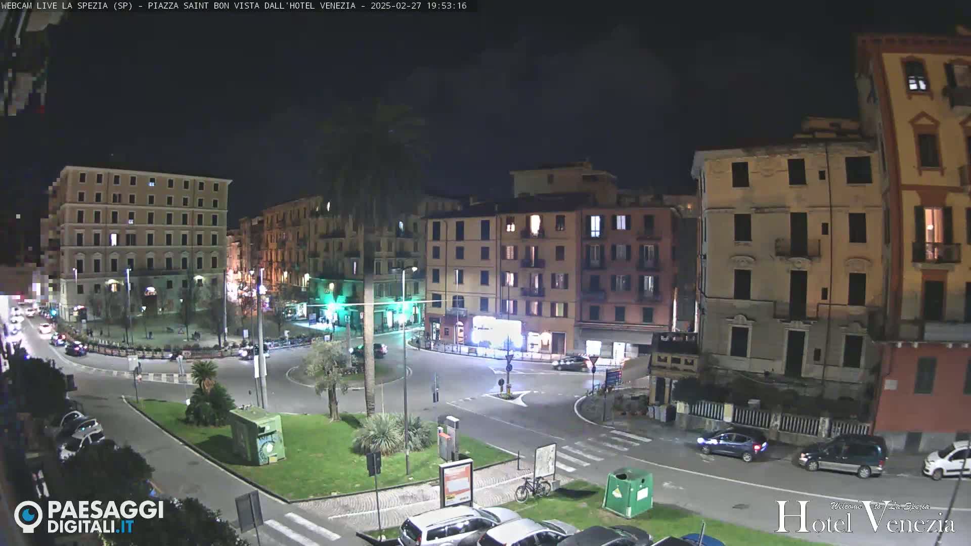 A nighttime view of a city square with several multi-story buildings, a small park, and sparse car traffic.