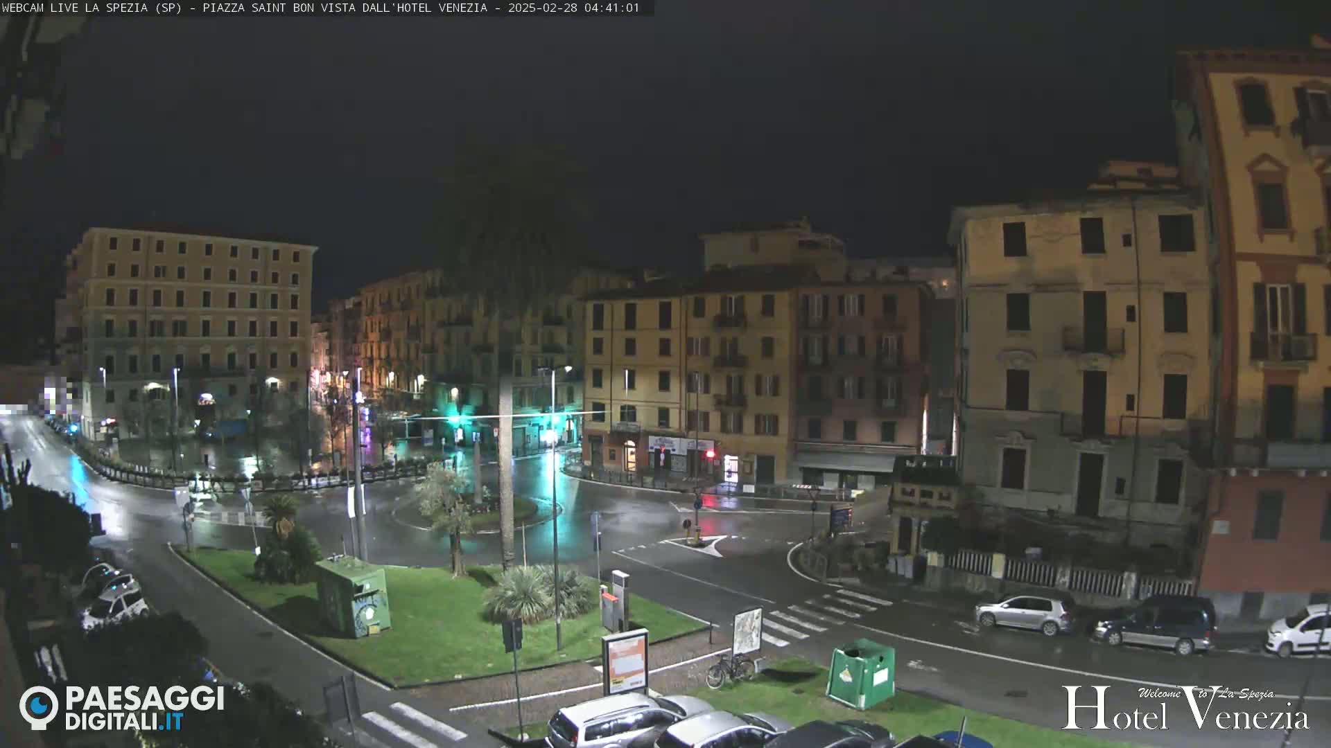 A nighttime, rain-slicked city square is surrounded by multi-story buildings, with cars parked along the perimeter and a few cars moving within the square.
