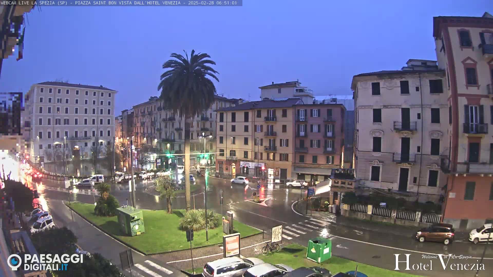 A rain-slicked town square in the early morning, surrounded by multi-story buildings, with a few cars and a palm tree visible.