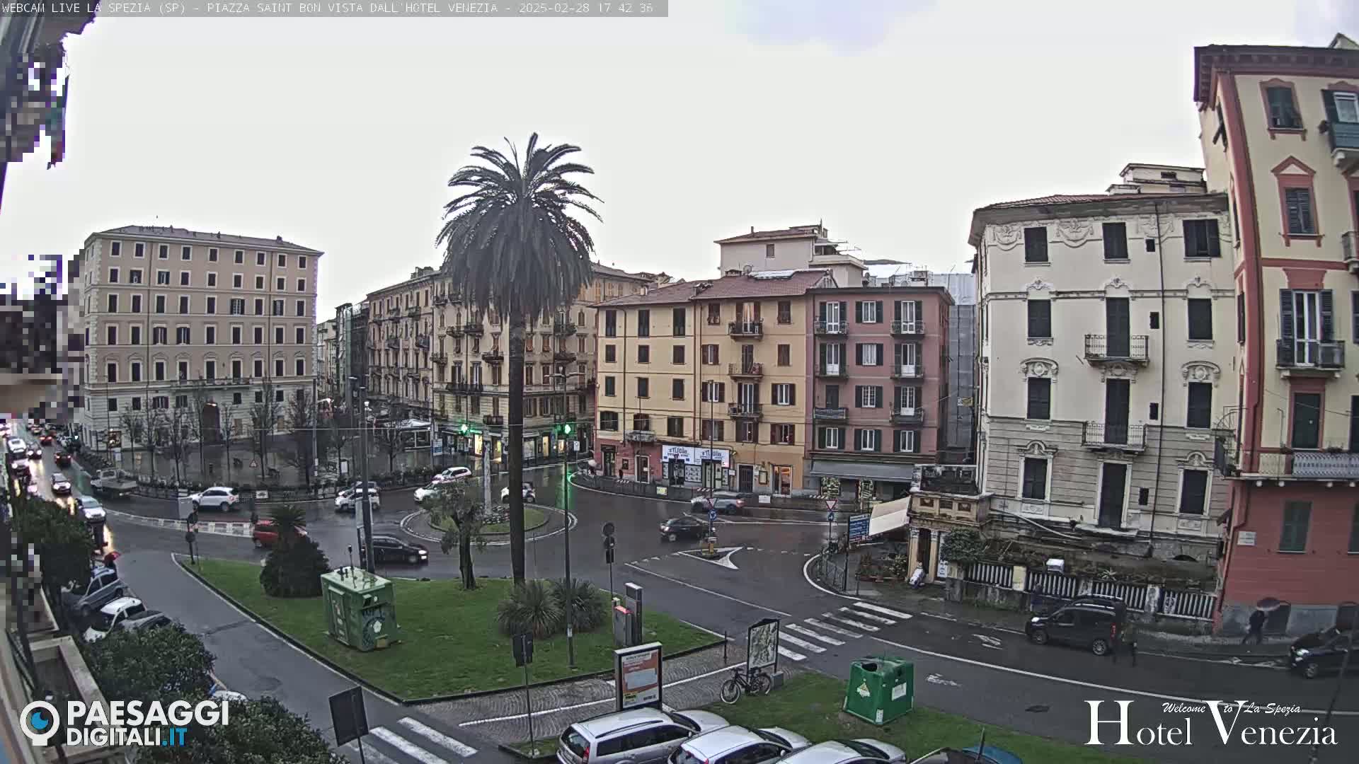 A wet, overcast city square with several multi-story buildings surrounding a roundabout where cars are driving.