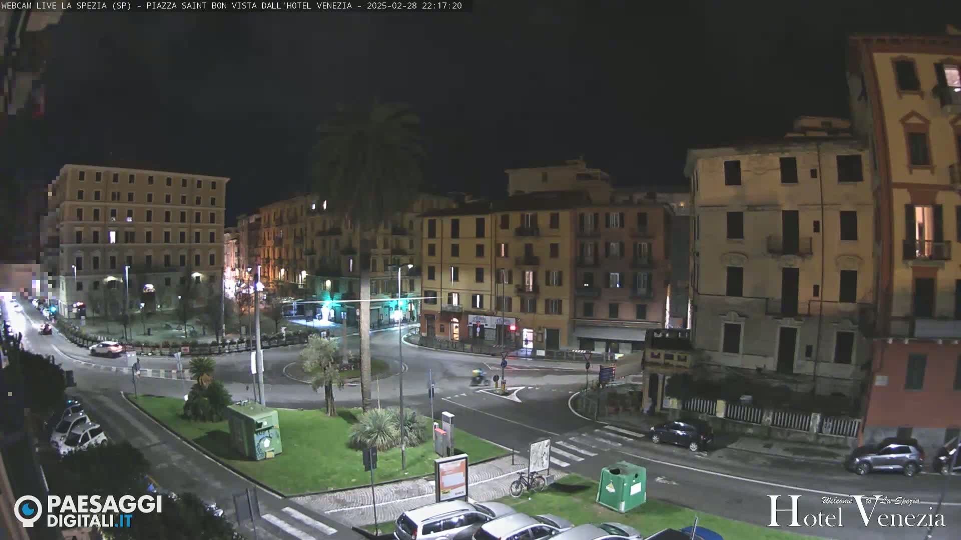 A nighttime, high-angle view of a town square in a European city, showing several multi-story buildings and cars parked along the perimeter, with a small amount of traffic on the roads.