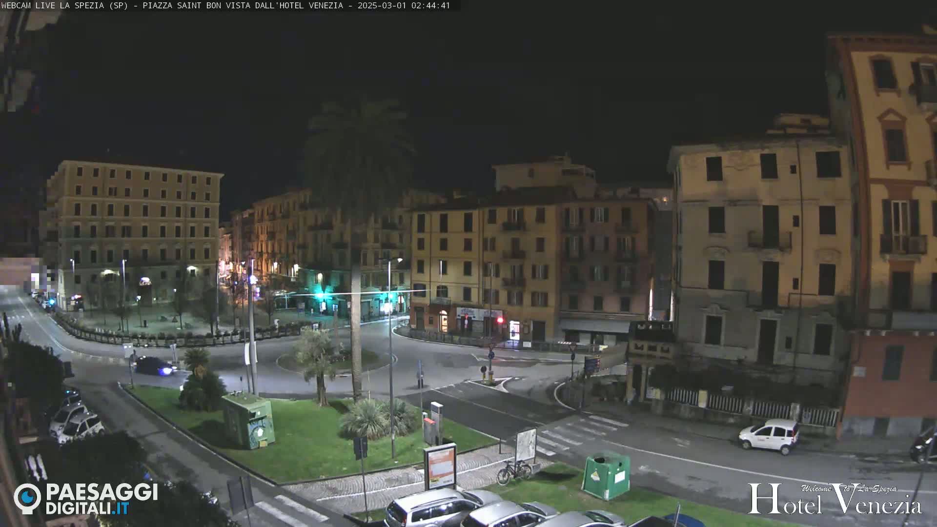 A nighttime view of a town square with several multi-story buildings surrounding a circular road, sparsely populated with vehicles.