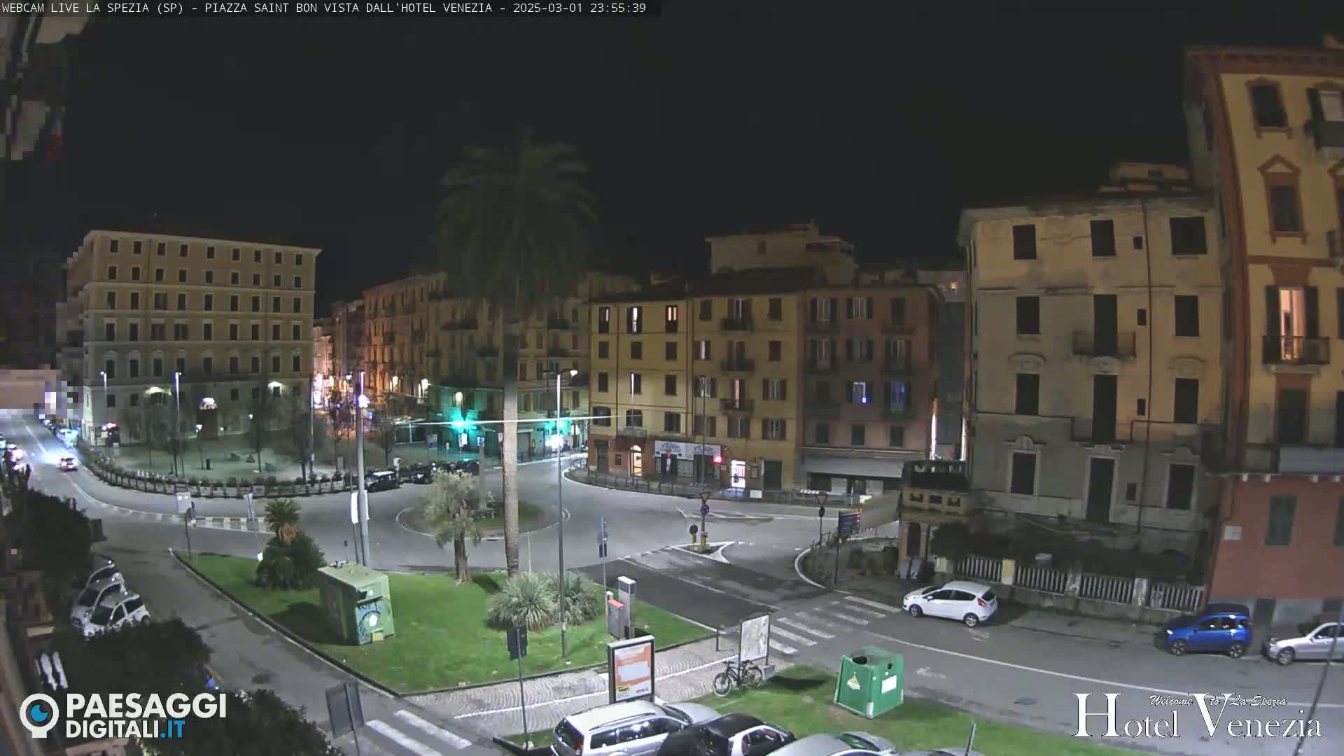 A nighttime view of a traffic circle in a European town square, surrounded by multi-story buildings under a clear, dark sky.
