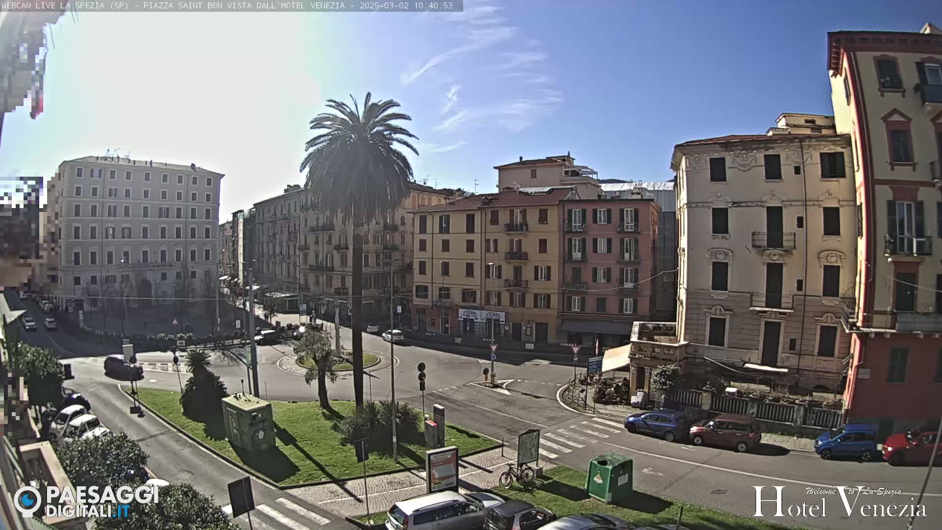 A sunny day reveals a traffic circle in a European city square, surrounded by multi-story buildings and sparse vehicle traffic.
