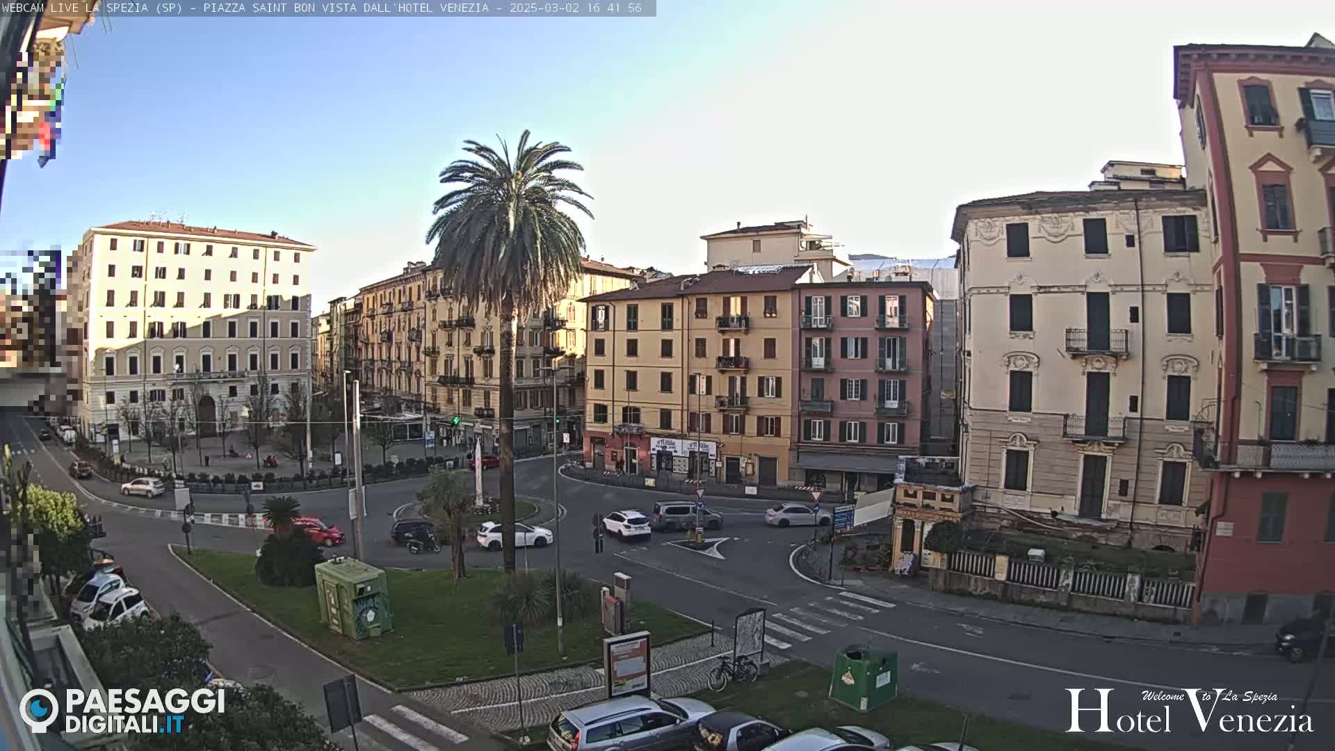 A mostly sunny day reveals a European city square with several multi-story buildings surrounding a roundabout with light traffic.
