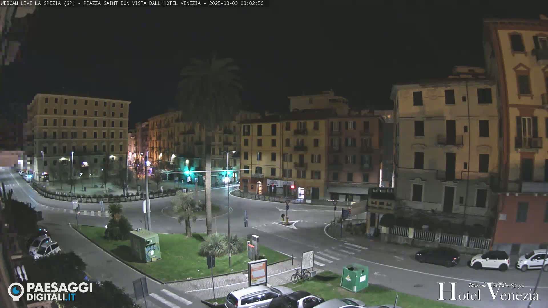 A nighttime view of a mostly empty town square surrounded by multi-story buildings, with a few cars parked and a bicycle present.