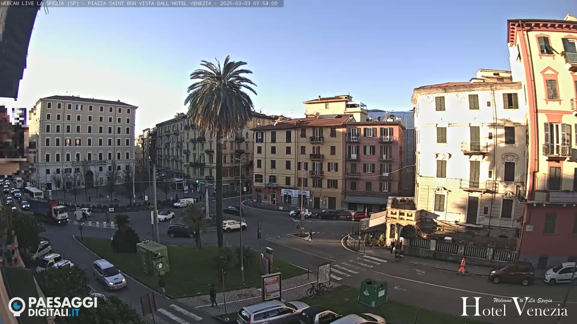 A sunny day in a European city square, showing several multi-story buildings, a palm tree in the center, cars parked and driving around a roundabout, and a few pedestrians walking.