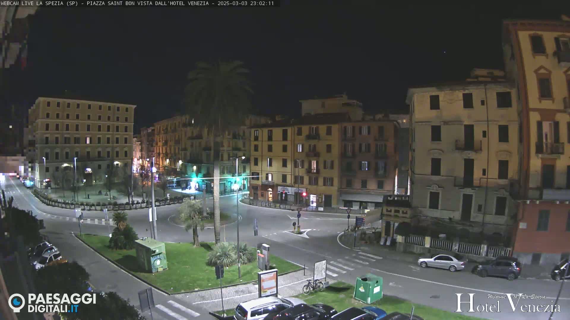 A nighttime view of a plaza in a city, surrounded by multi-story buildings, with a few cars parked and a bicycle visible.
