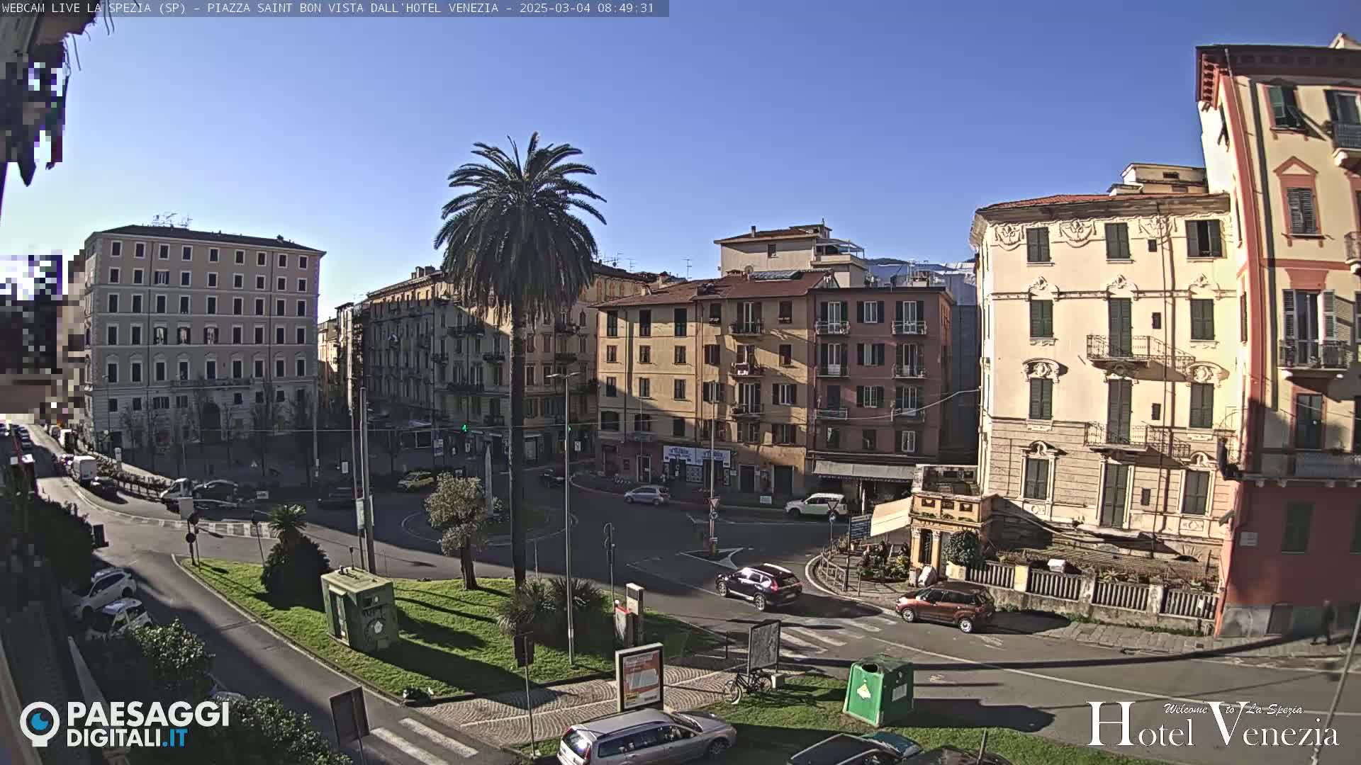 A sunny day in a European city square, showing several multi-story buildings and cars driving around a central, tree-filled circle.