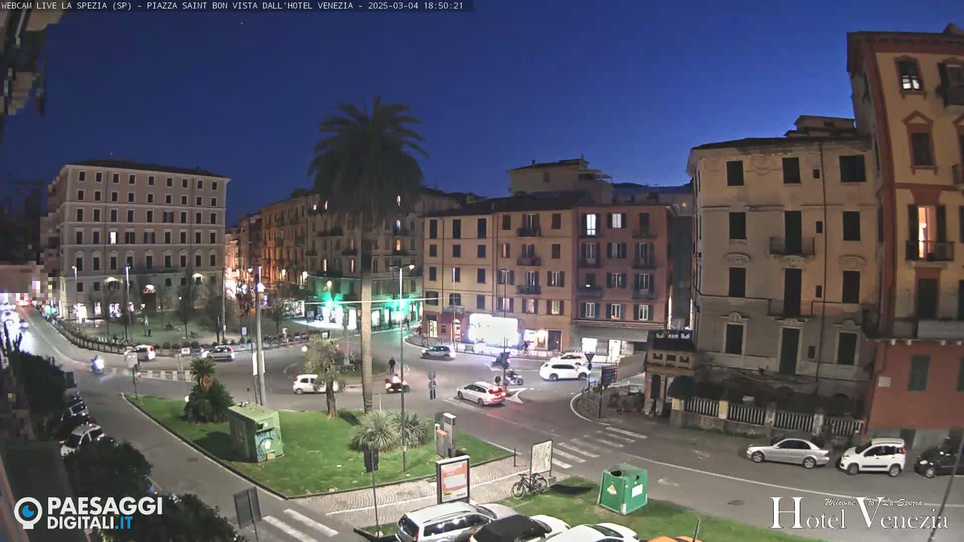 A nighttime view of a town square with several multi-story buildings, some cars, and a palm tree, under a clear, dark blue sky.