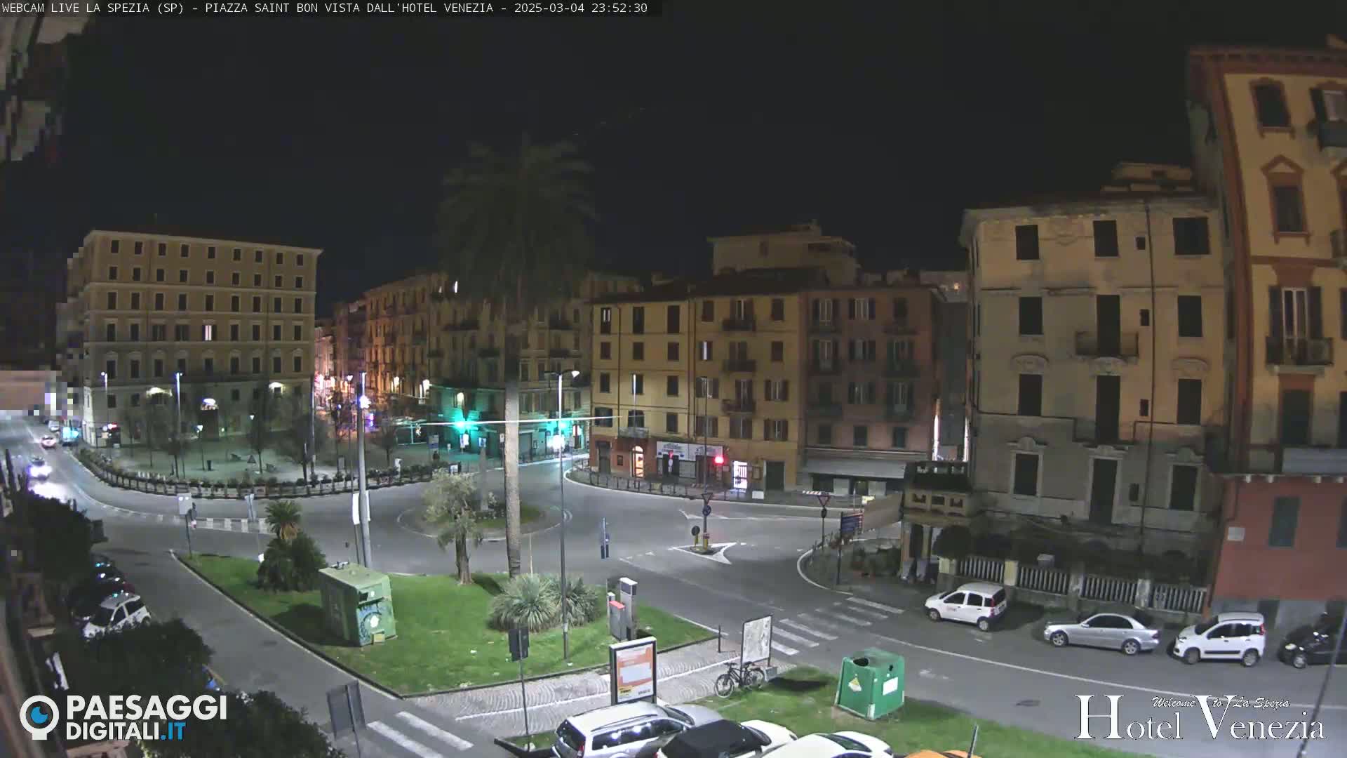 A nighttime view of a town square with several multi-story buildings surrounding it, illuminated by streetlights, with cars parked along the perimeter and a few driving through the square.