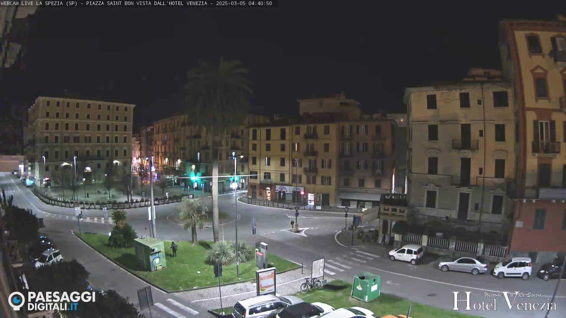 A nighttime, high-angle view of a mostly empty plaza surrounded by multi-story buildings, with a few cars and a bicycle visible.
