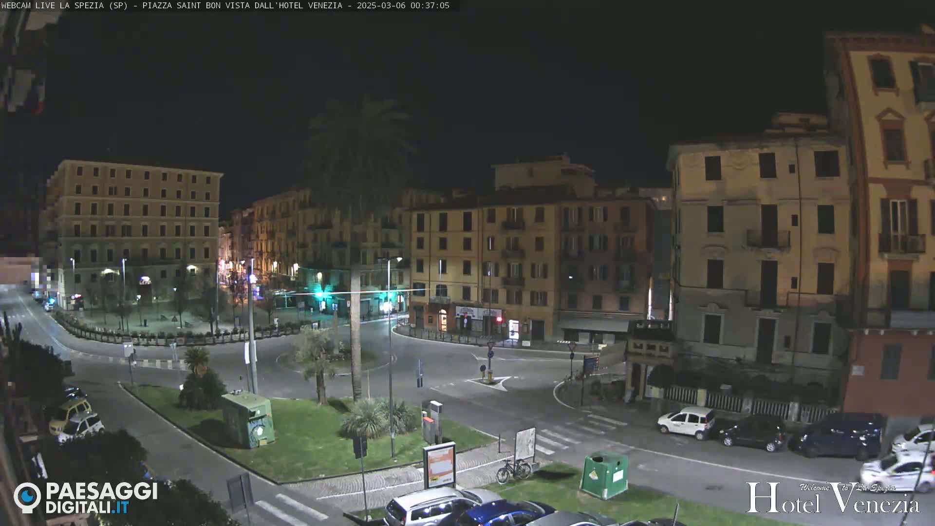 A nighttime view of a quiet town square in Italy, surrounded by multi-story buildings, with a few cars parked and a bicycle visible.