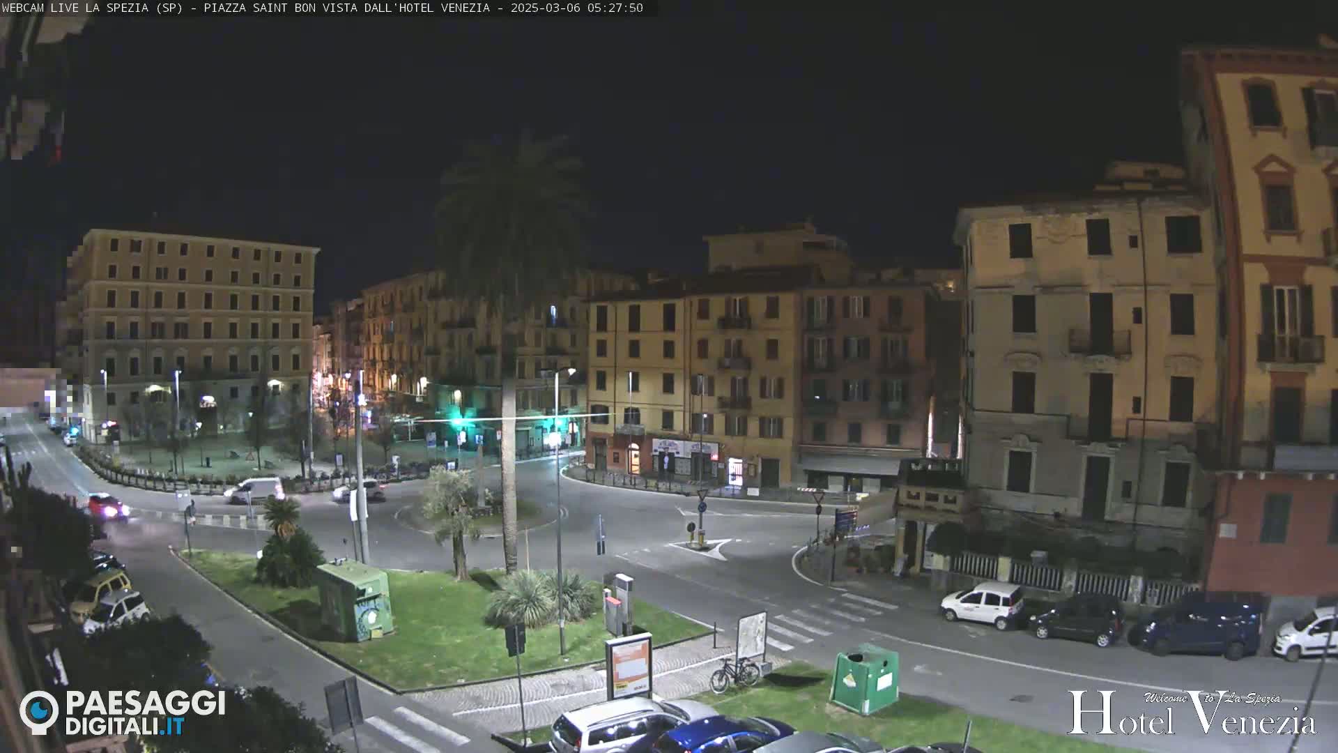 A nighttime view of a city square with several multi-story buildings, some cars, and a palm tree, under dark, clear skies.