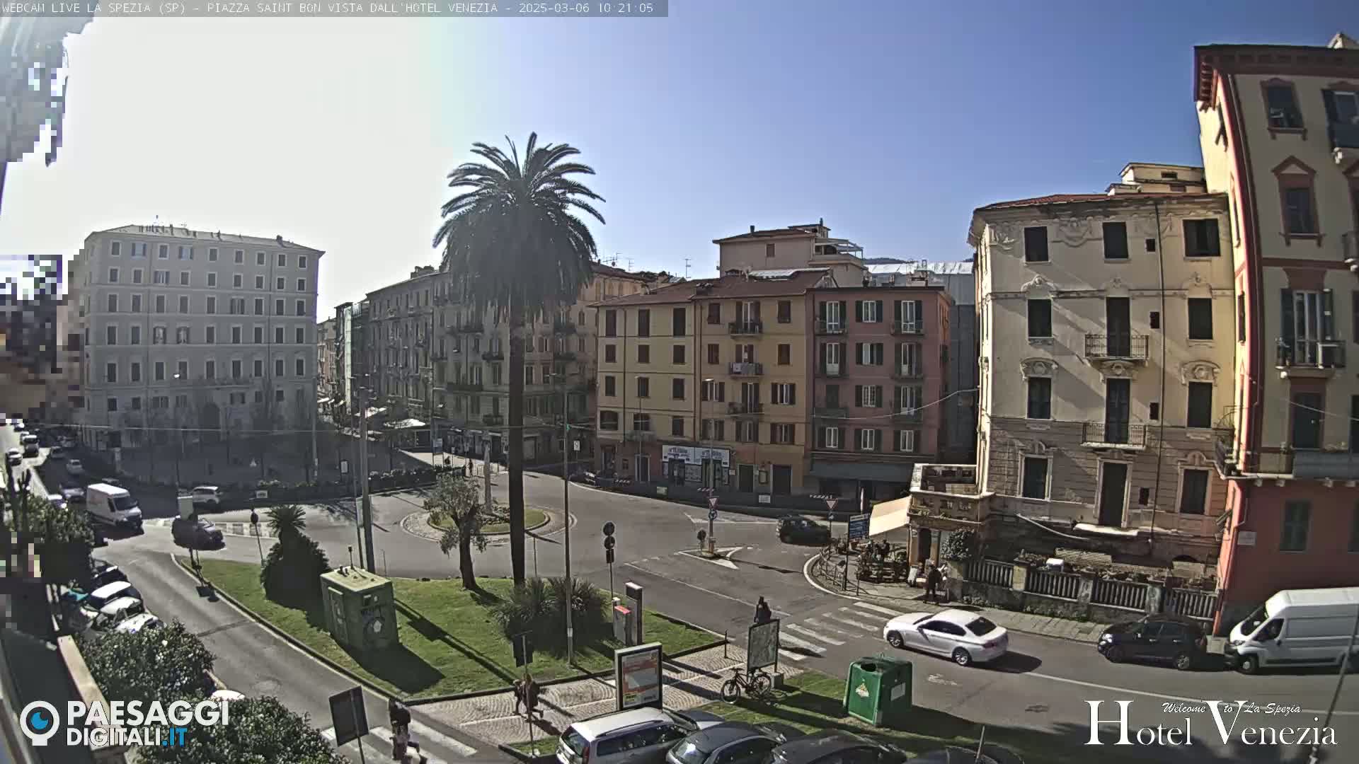A sunny day reveals a European city square with several multi-story buildings surrounding a small roundabout, where cars and a few pedestrians are visible.