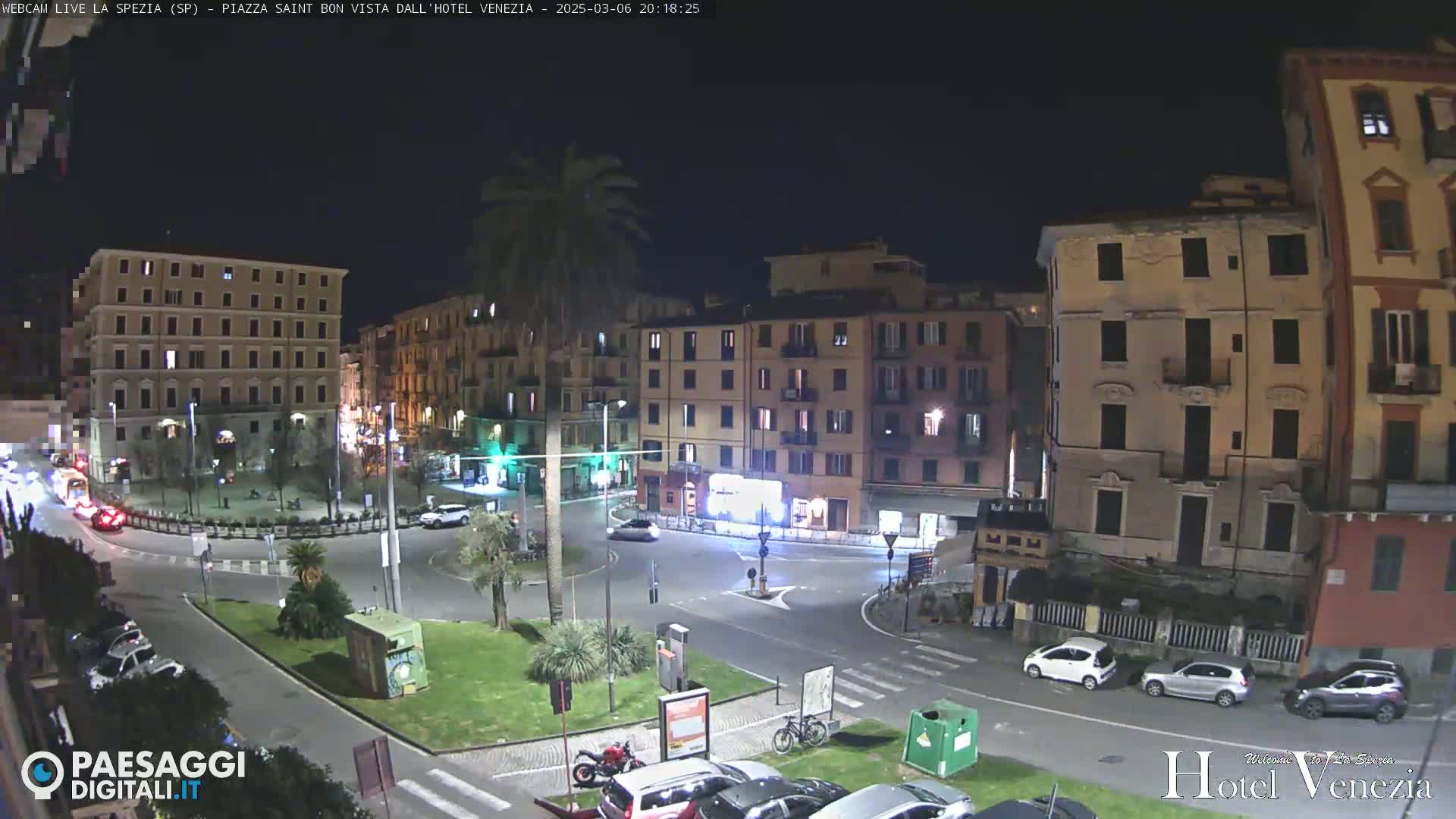 A nighttime view of a city square with several multi-story buildings surrounding a roundabout, where a few cars are visible.