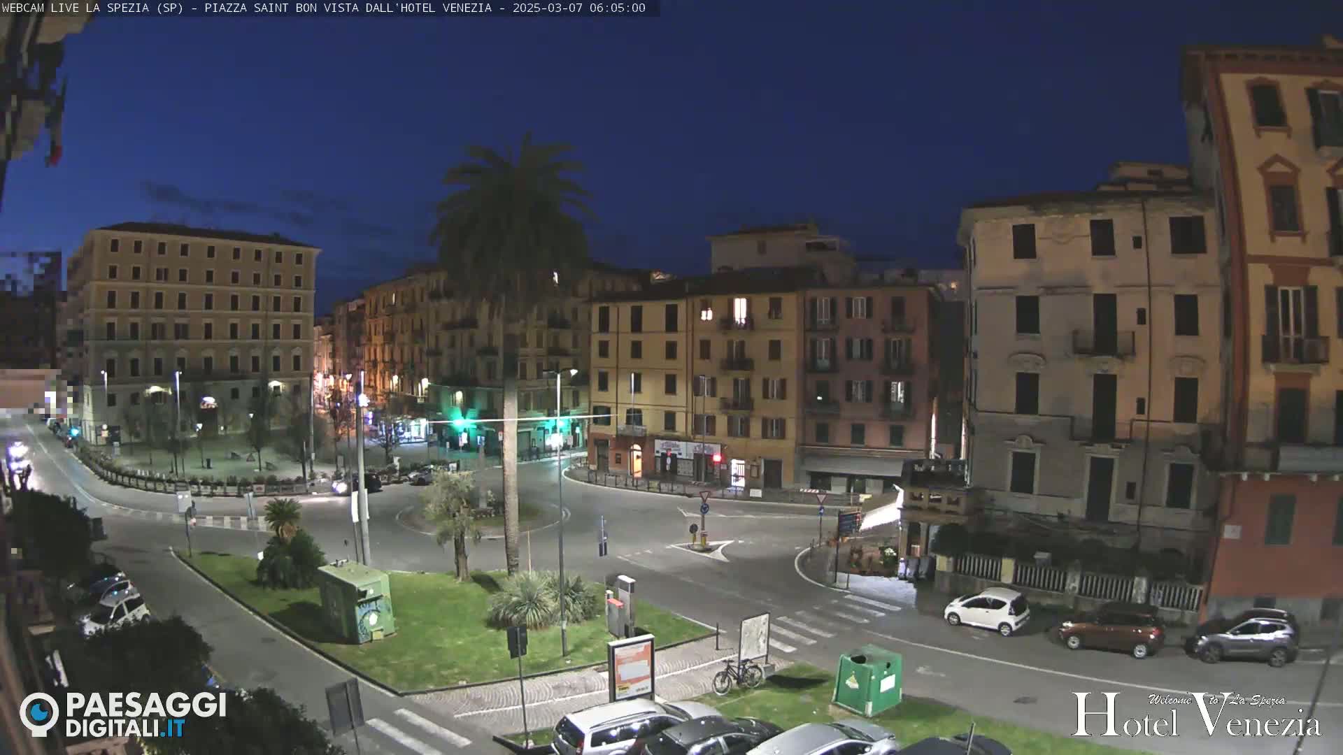 A nighttime view of a European city square, with several multi-story buildings illuminated, and cars parked and driving around a circular traffic area.