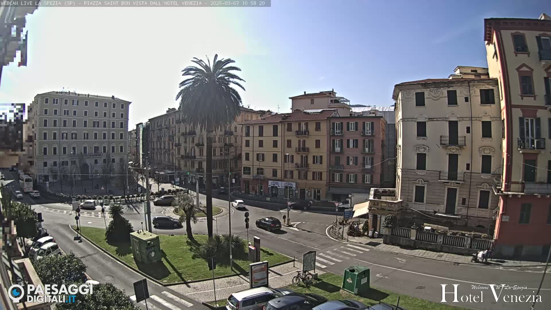 A sunny day in a European city square, showing multi-story buildings surrounding a circular roadway with sparse traffic and a central palm tree.