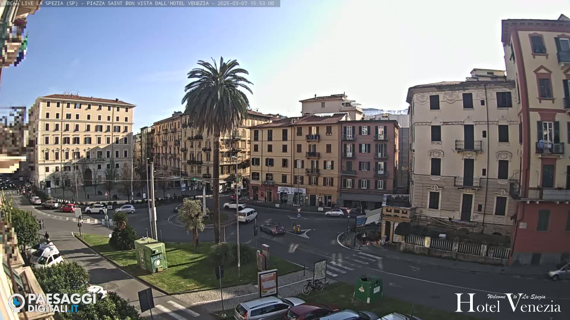 A sunny day overlooks a European-style city square with several multi-story buildings, palm trees, and cars moving around a roundabout.