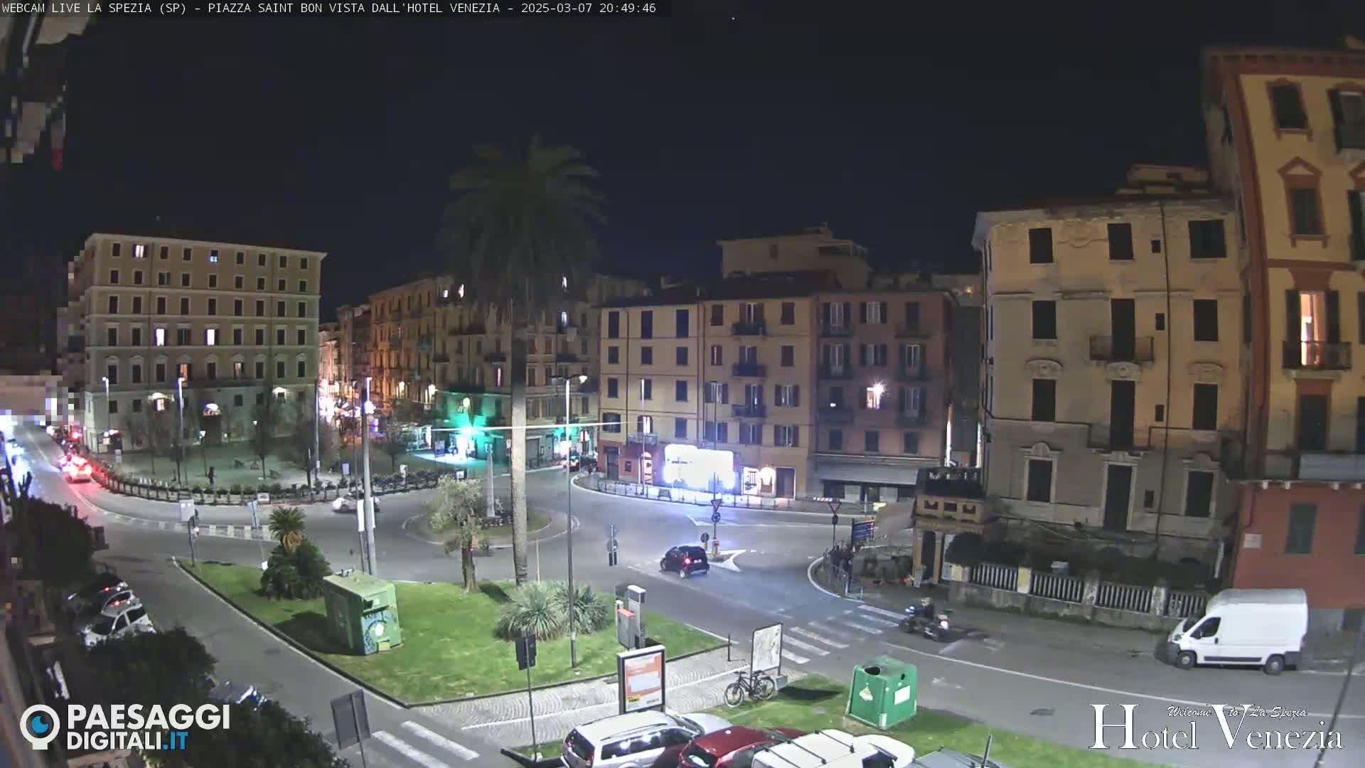 A nighttime view of a town square with several multi-story buildings surrounding a circular roadway, featuring sparse vehicle traffic and a few pedestrians.