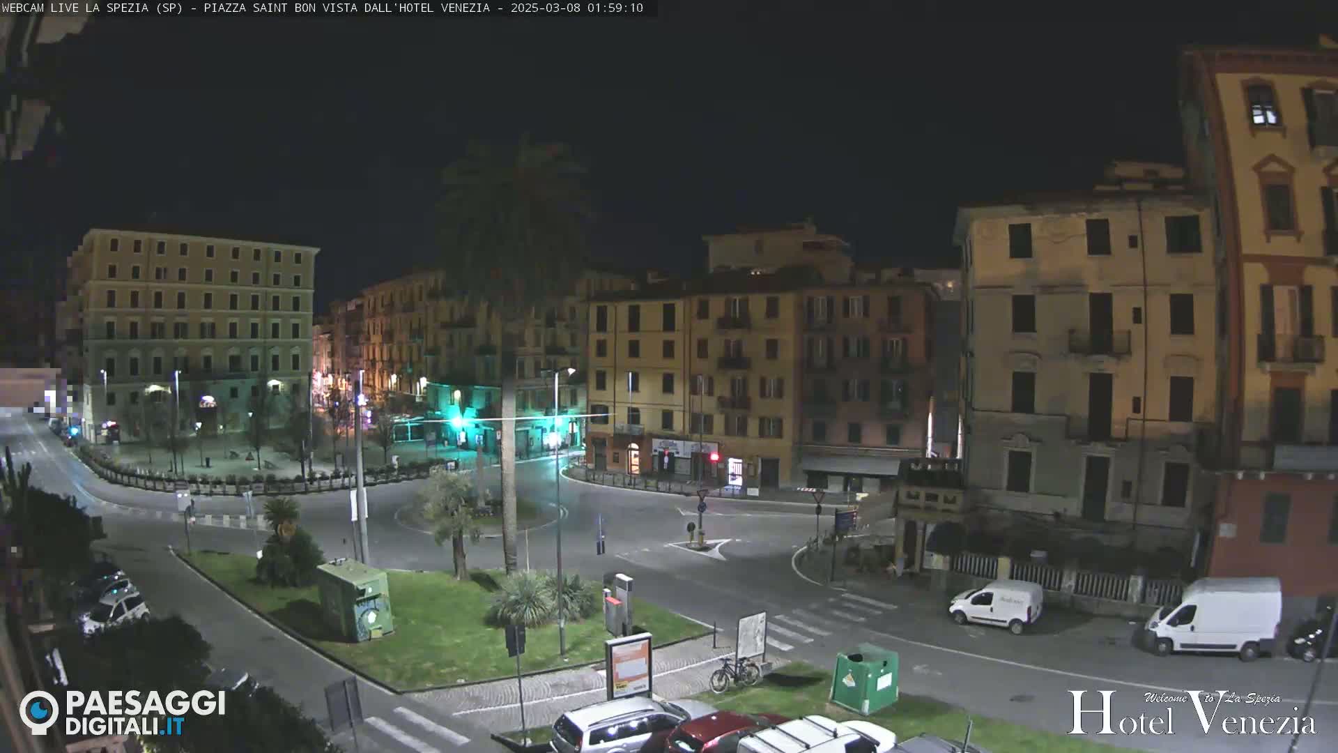 A nighttime view of a mostly empty city square surrounded by multi-story buildings, with a few cars and a bicycle visible.