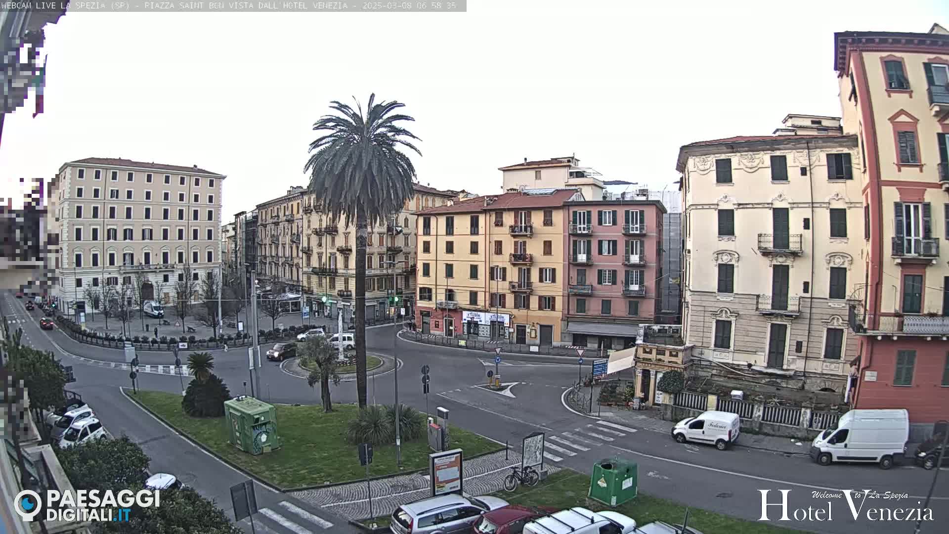 A mostly overcast view of a town square with several multi-story buildings, a roundabout, parked cars, and a palm tree in the center.