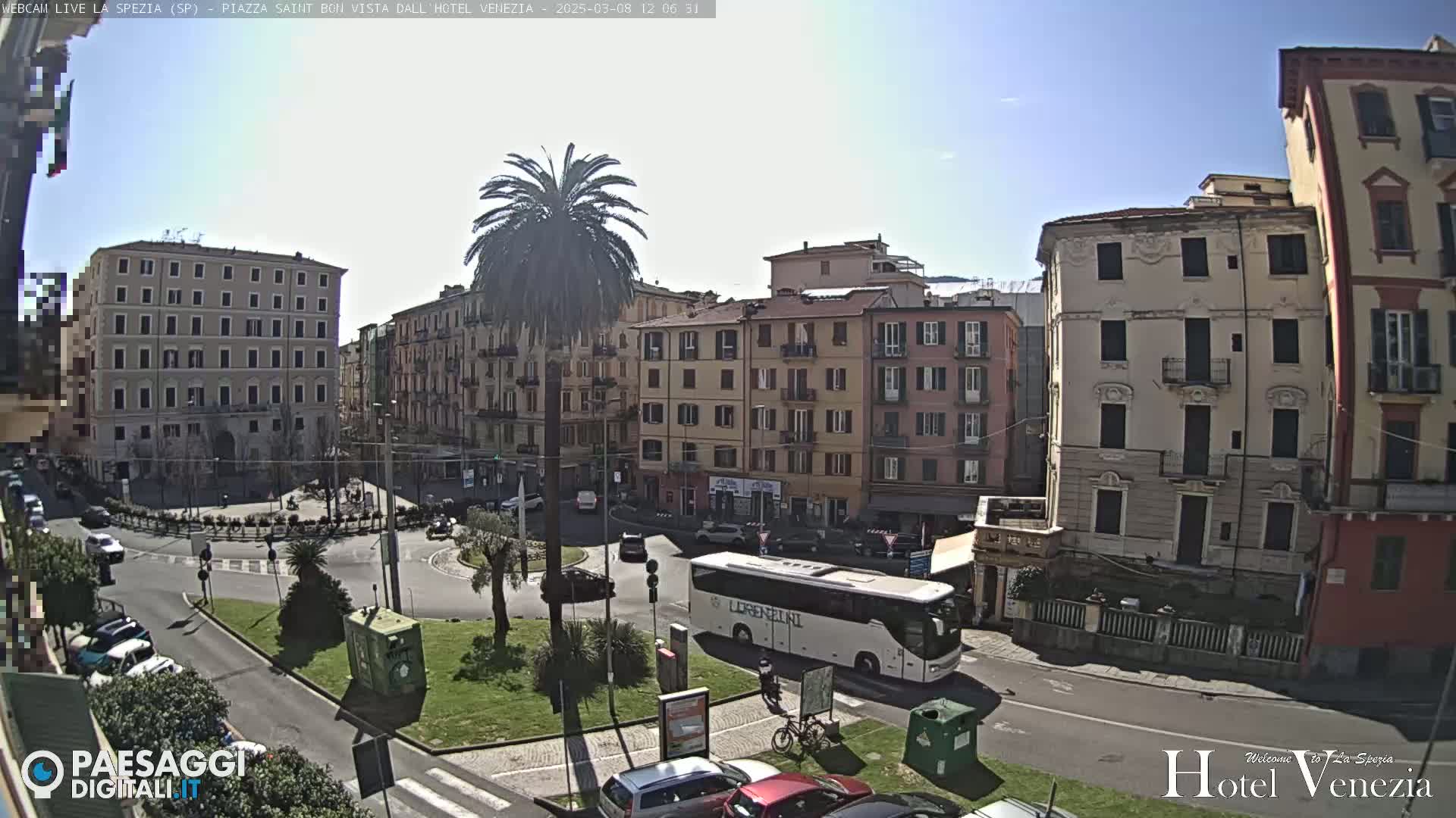 A sunny day in a European city square, showing several multi-story buildings, a palm tree, cars, and a large tour bus.