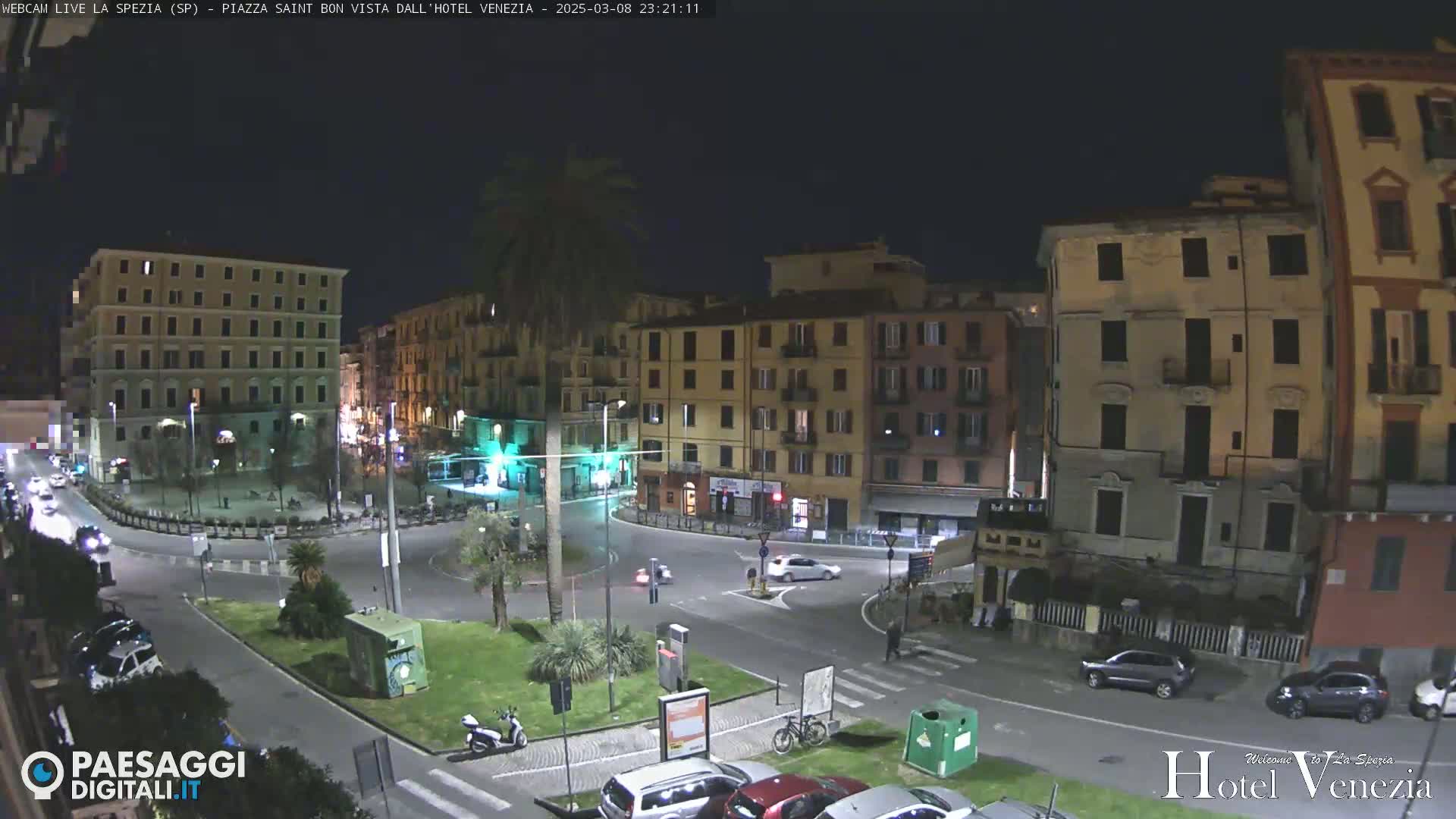 A nighttime view of a city square with several buildings surrounding a circular roadway, with sparse vehicle traffic and a few pedestrians.