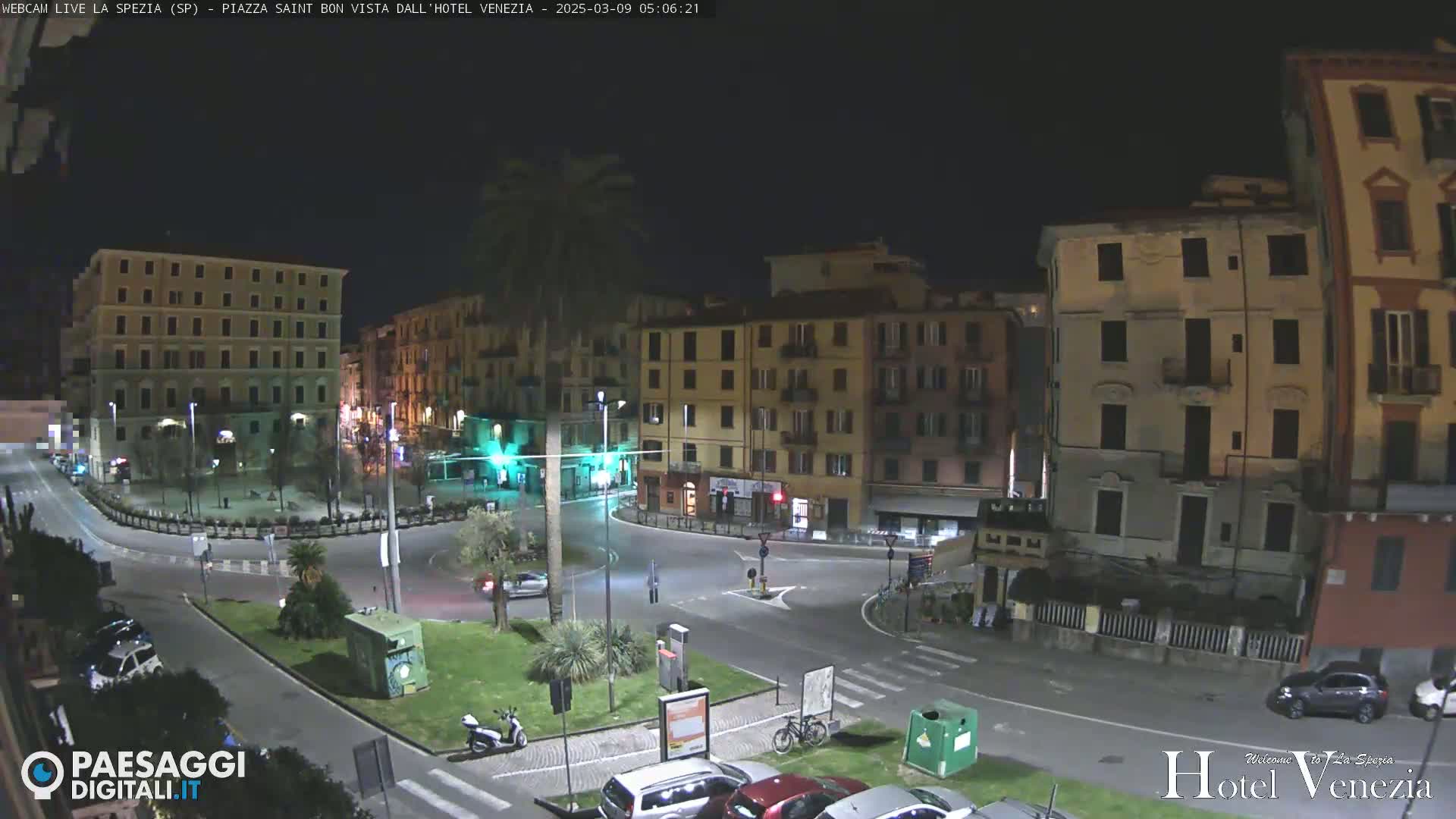 A nighttime view of a mostly empty town square surrounded by multi-story buildings, with a few cars and a scooter visible.