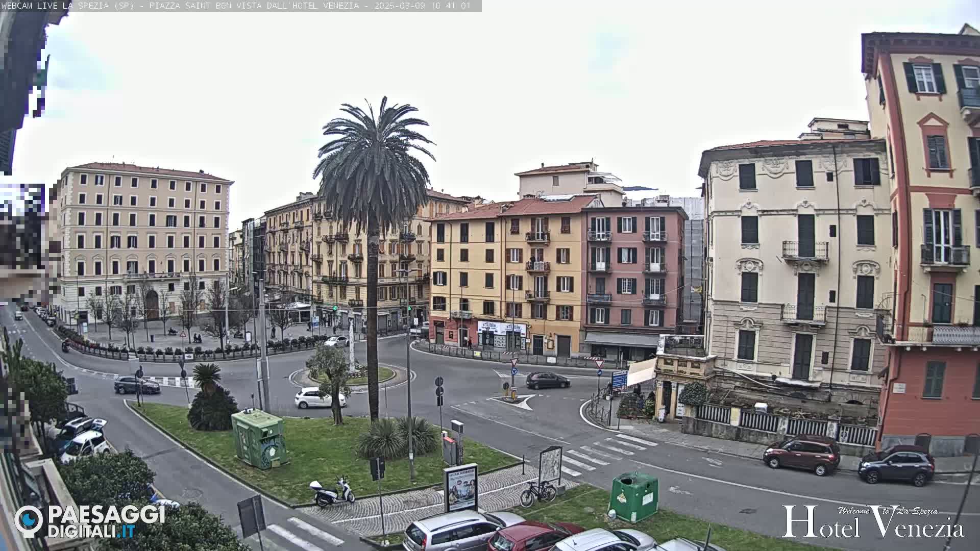 A mostly cloudy day in a European city square, with several multi-story buildings surrounding a roundabout where cars and a scooter are visible.