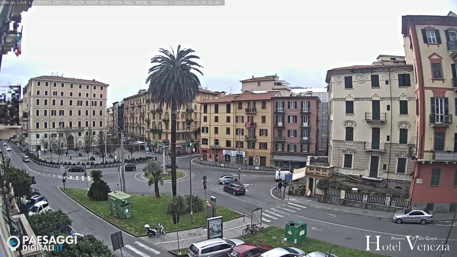 A mostly cloudy day in a European city square, where several multi-story buildings surround a roundabout with sparse traffic and a lone palm tree.
