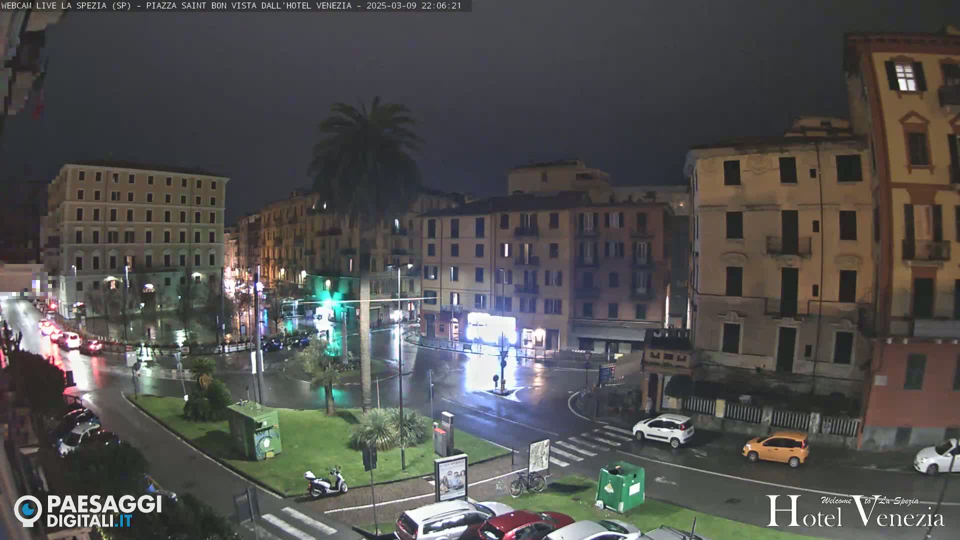 A nighttime view of a rain-slicked city square surrounded by multi-story buildings, with cars and a scooter visible.