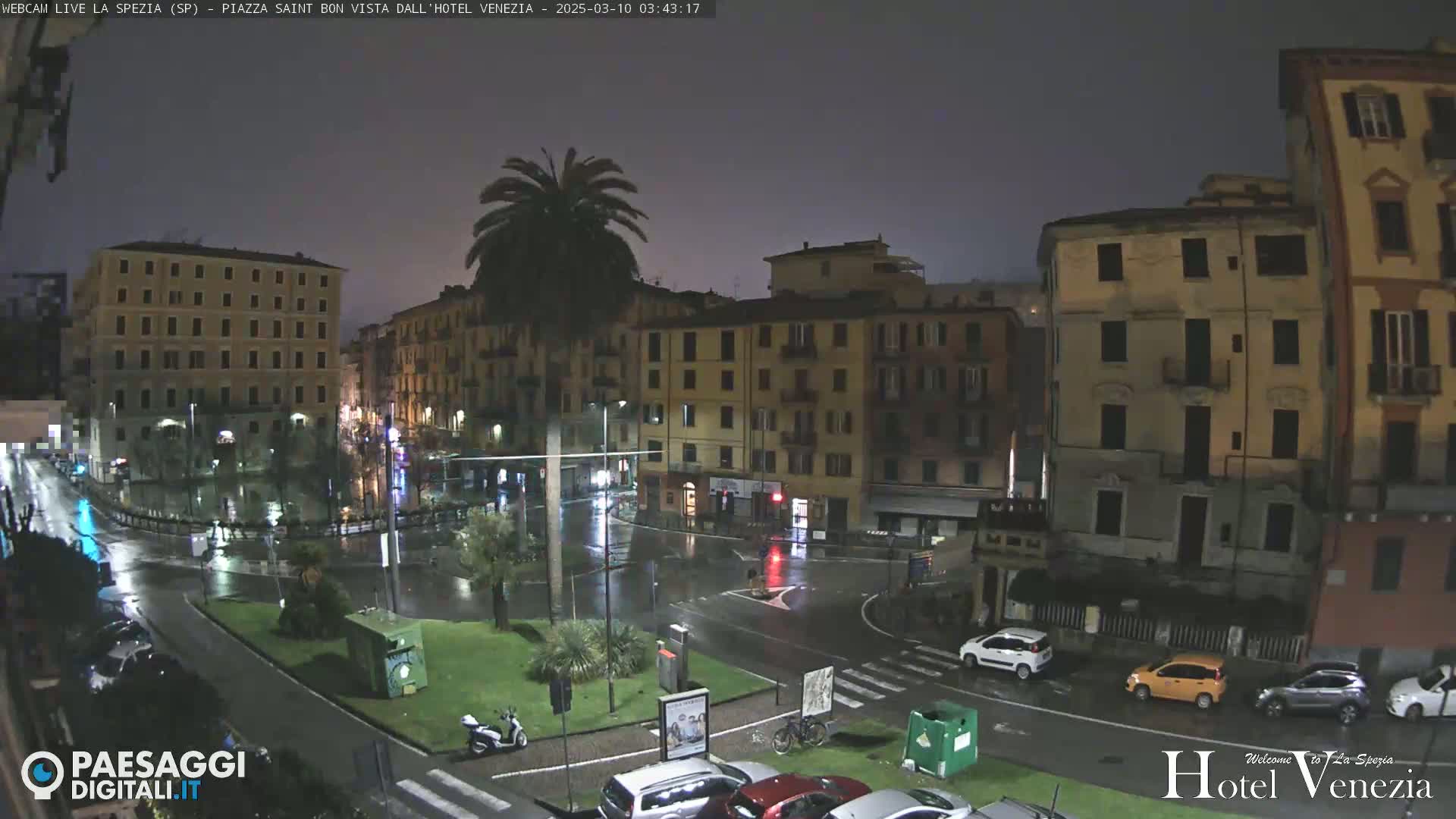 A rain-slicked plaza at night, surrounded by multi-story buildings, with a few cars and a scooter parked.