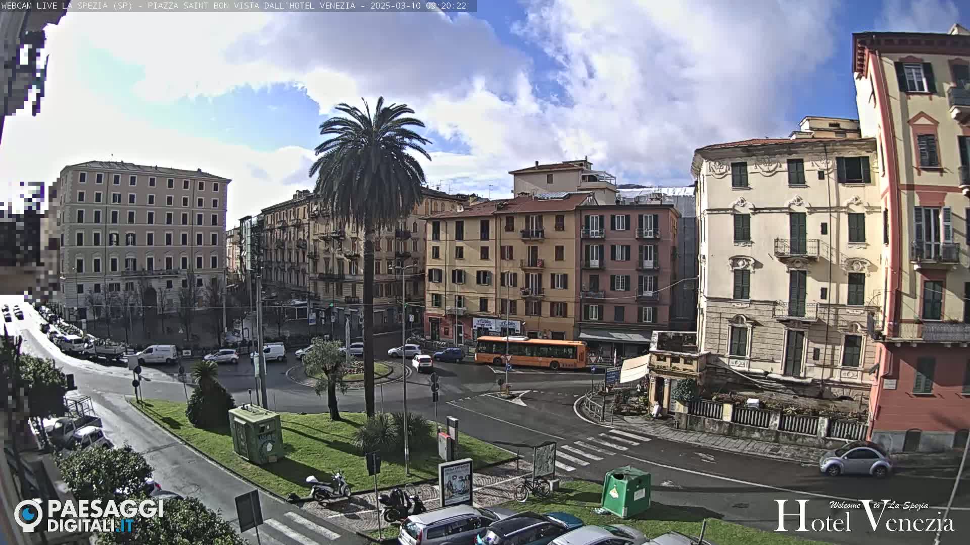 A partly cloudy day reveals a city square with several multi-story buildings, a palm tree, and light traffic.