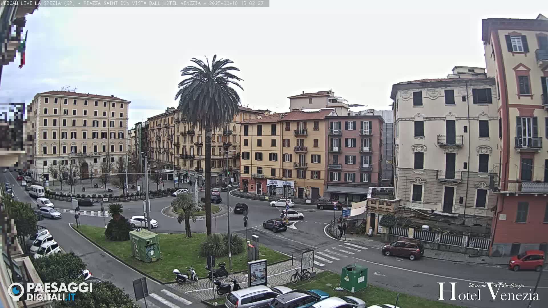 A mostly cloudy day reveals a city square with several multi-story buildings, a palm tree in a roundabout, and several cars.