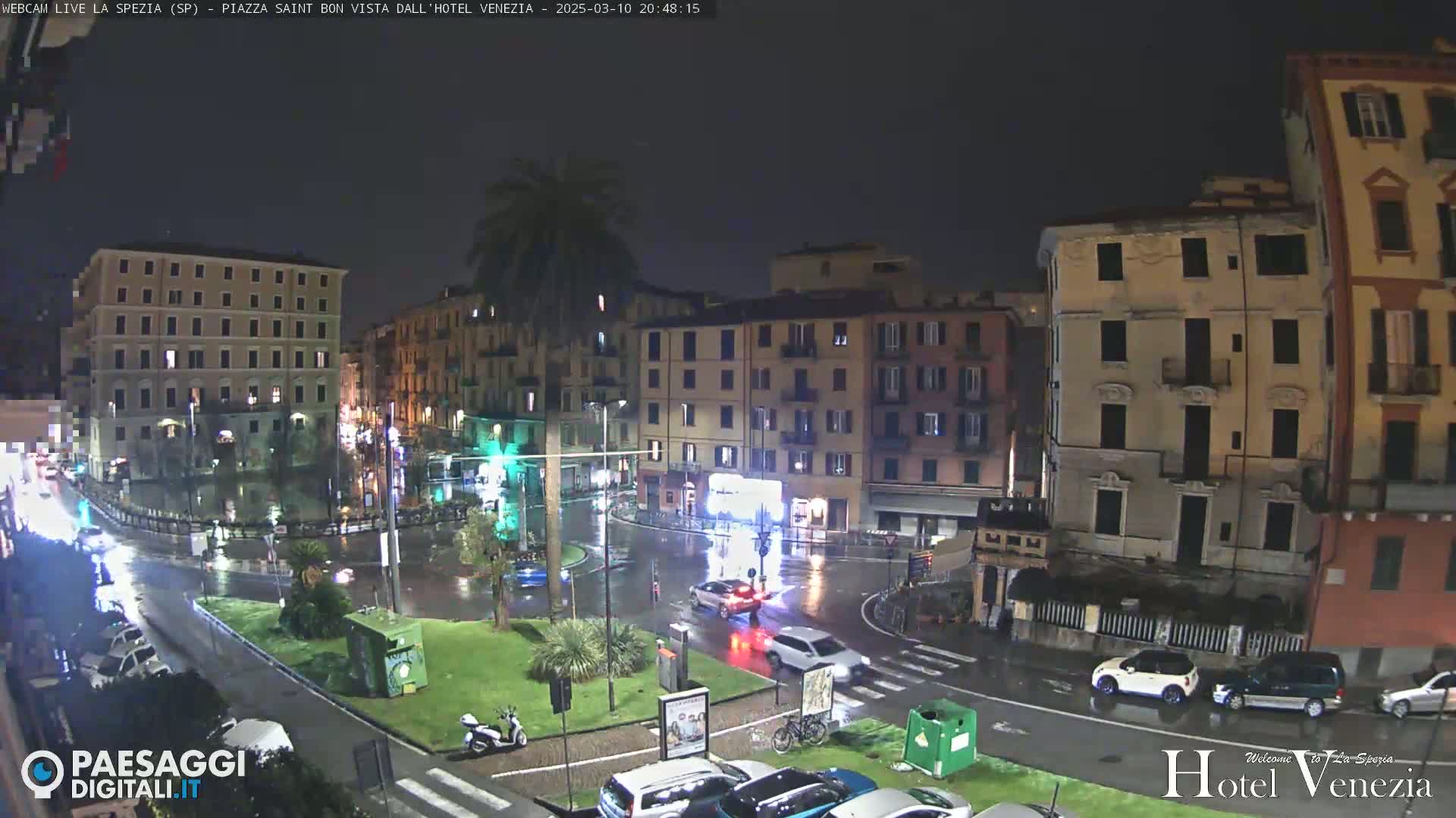 A nighttime, rainy view of a city square surrounded by multi-story buildings, with cars and a scooter visible on wet streets.
